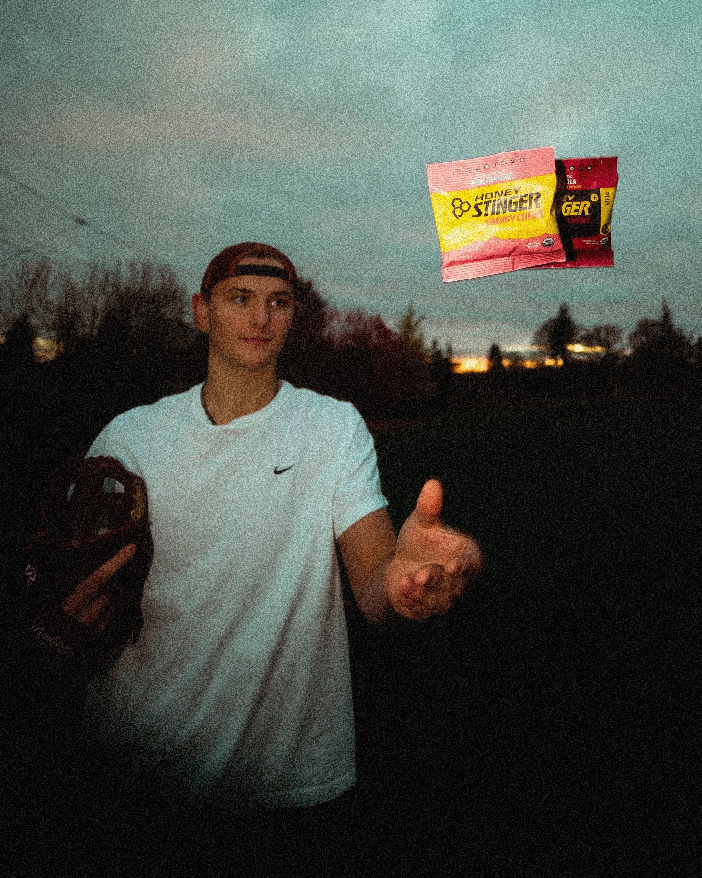 Young man wearing a white Nike shirt, baseball cap backwards, standing outdoors at sunset with trees and open sky in the background. He is holding a bag of Honey Stinger energy chews and gesturing with his hand.