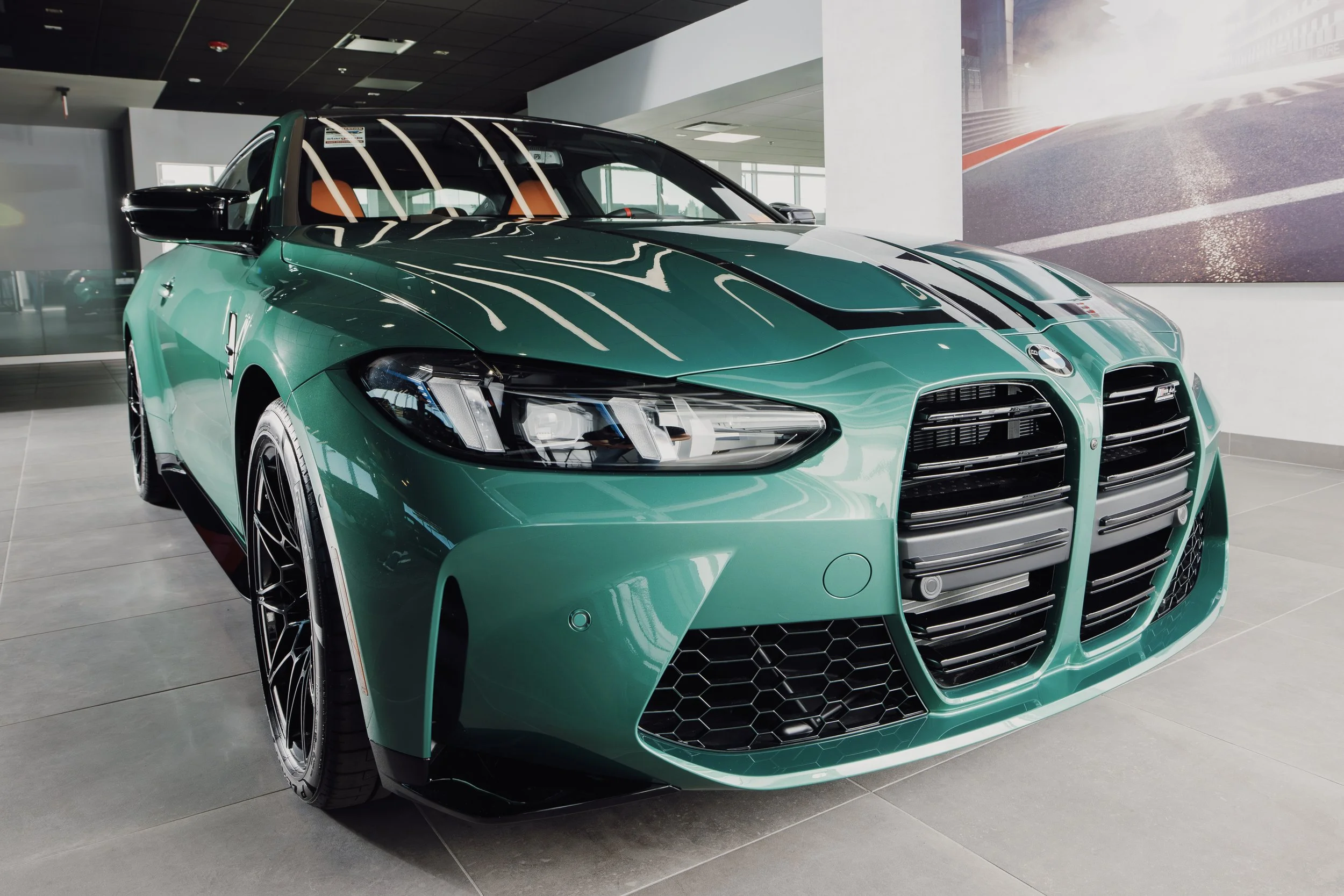 A teal-colored BMW M8 car displayed indoors at a showroom with a large mural of a race track in the background.