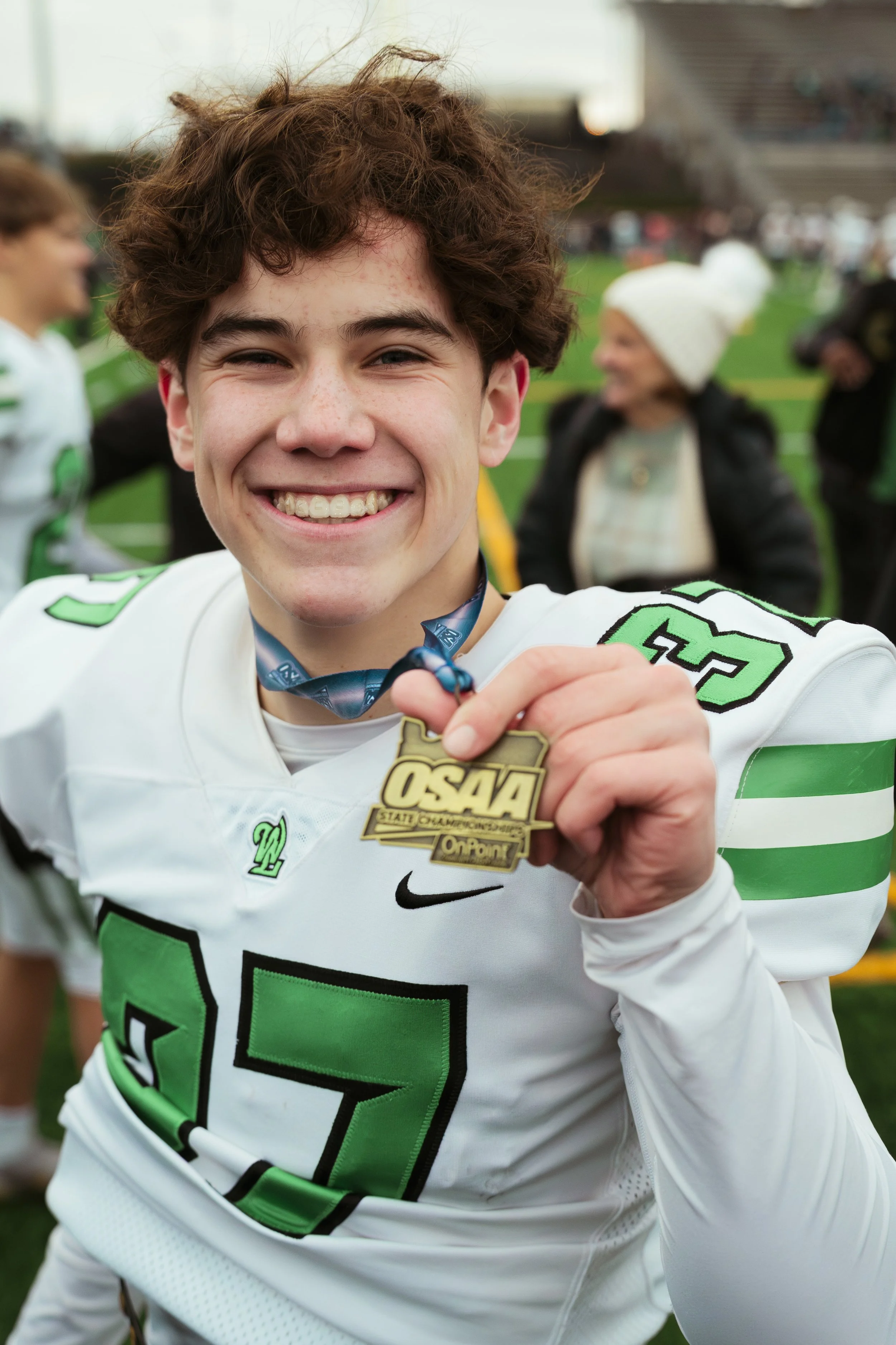 A young male athlete in a football uniform smiling and showing a medal, on a sports field with other people in the background.
