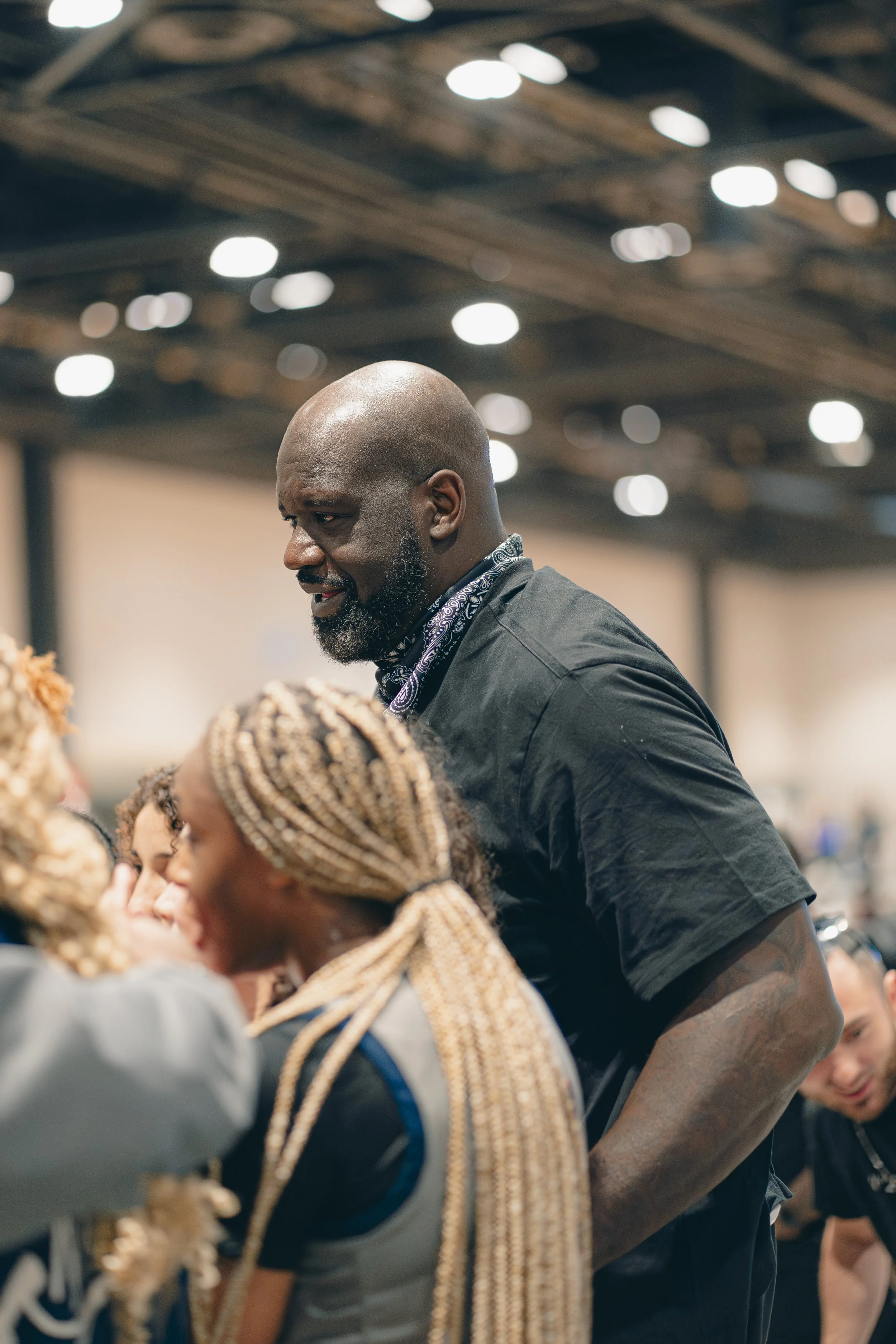 A man with a bald head, gray beard, wearing a black shirt with a bandana around his neck, is smiling while standing among a group of people with braided hair. The setting appears to be indoors with an industrial-style ceiling with lights.