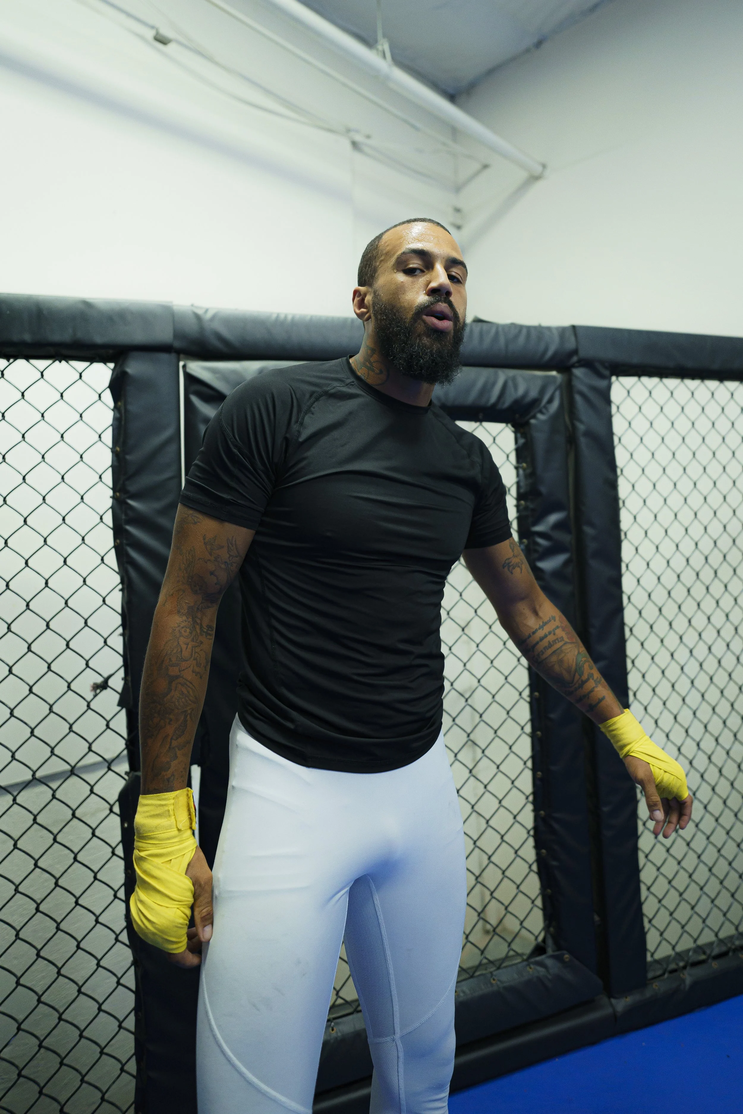 A male mixed martial artist standing inside a cage at a training facility, wearing black athletic shirt, white shorts, yellow gloves, and with tattoos on his arm and neck.