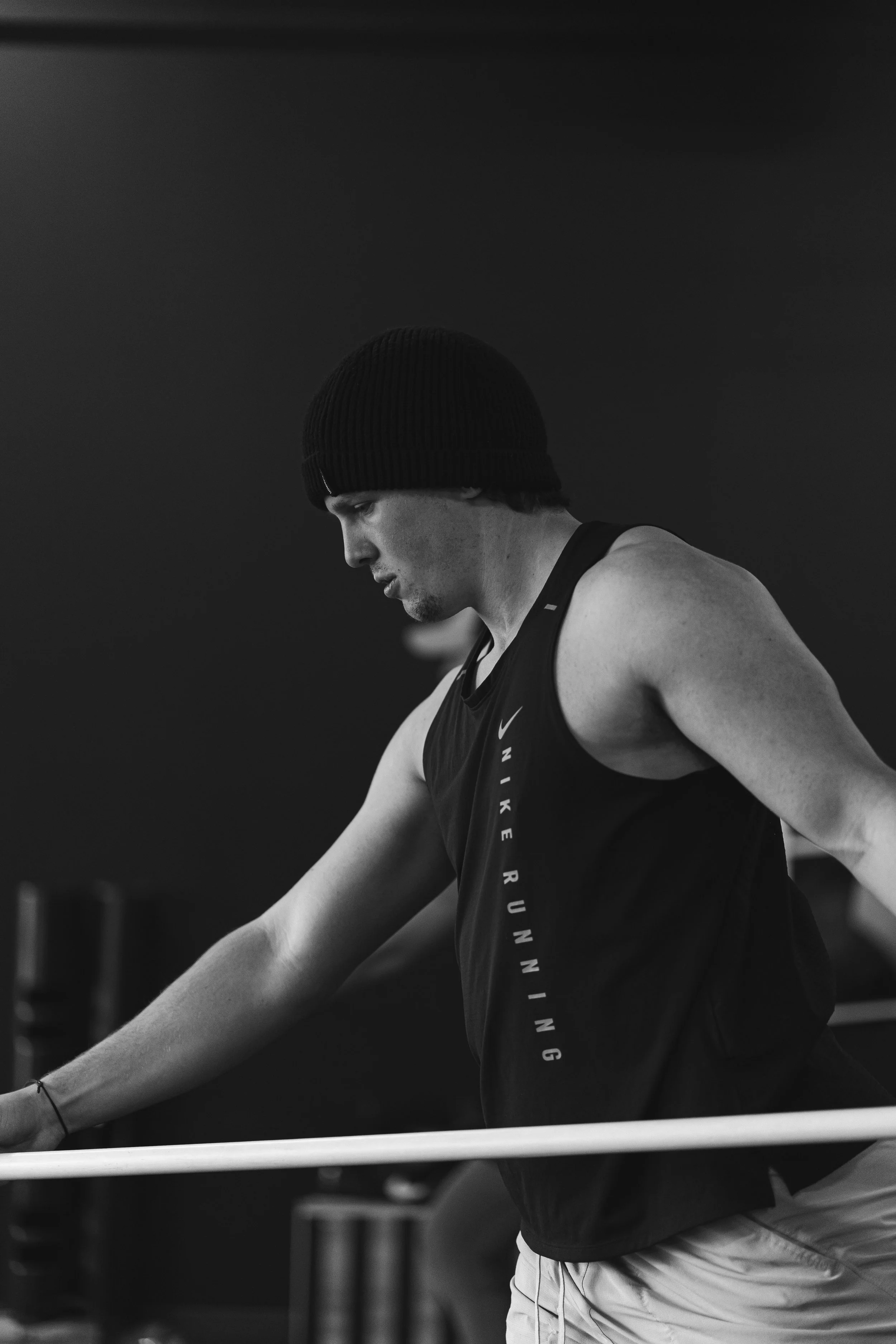 A young man wearing a knit cap and athletic wear is leaning on a railing, looking down thoughtfully, in a black and white photo.