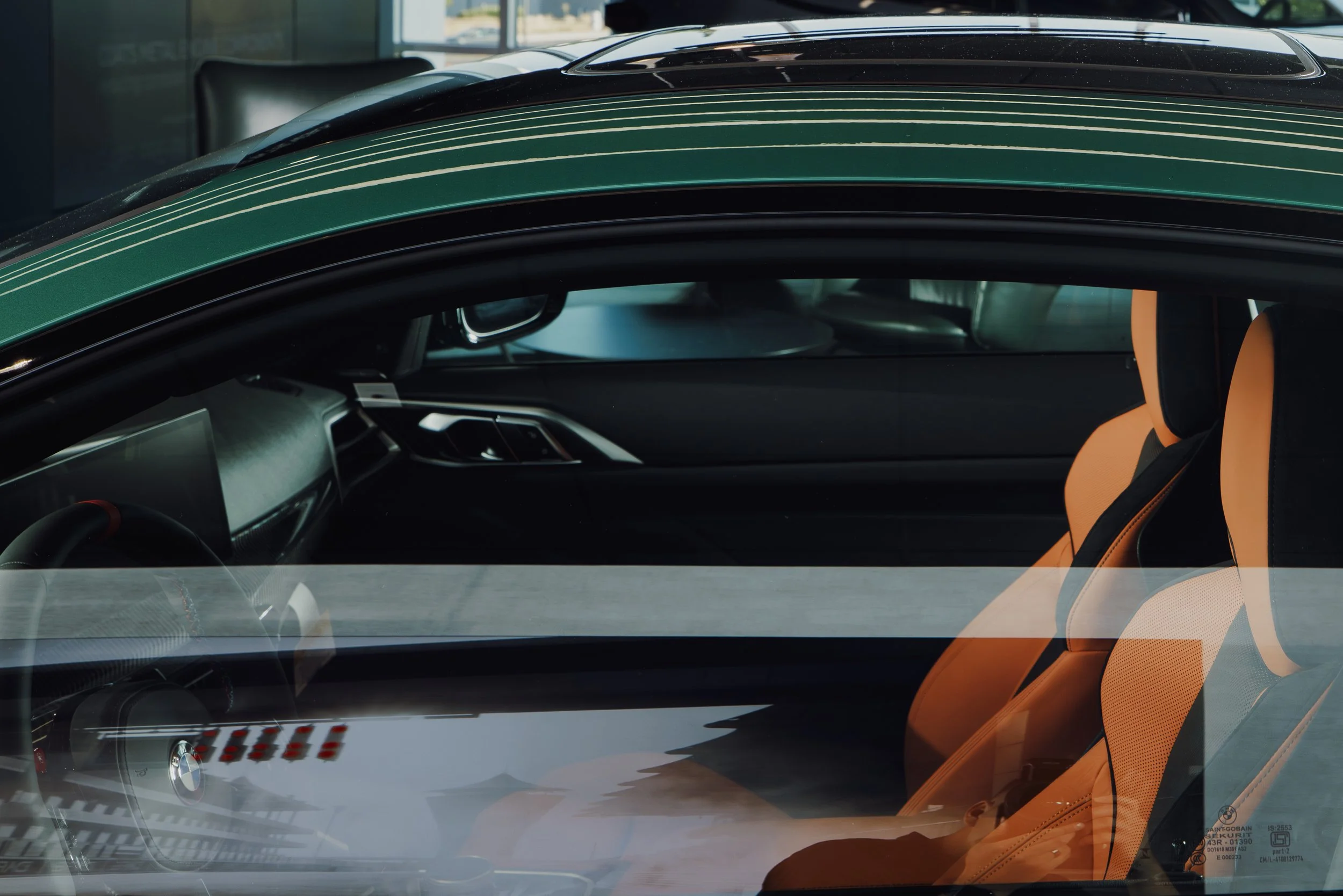 The image shows the interior of a luxury sports car, with a focus on the orange leather seats, steering wheel, and dashboard. The windshield reflects the ceiling lights. BMW.