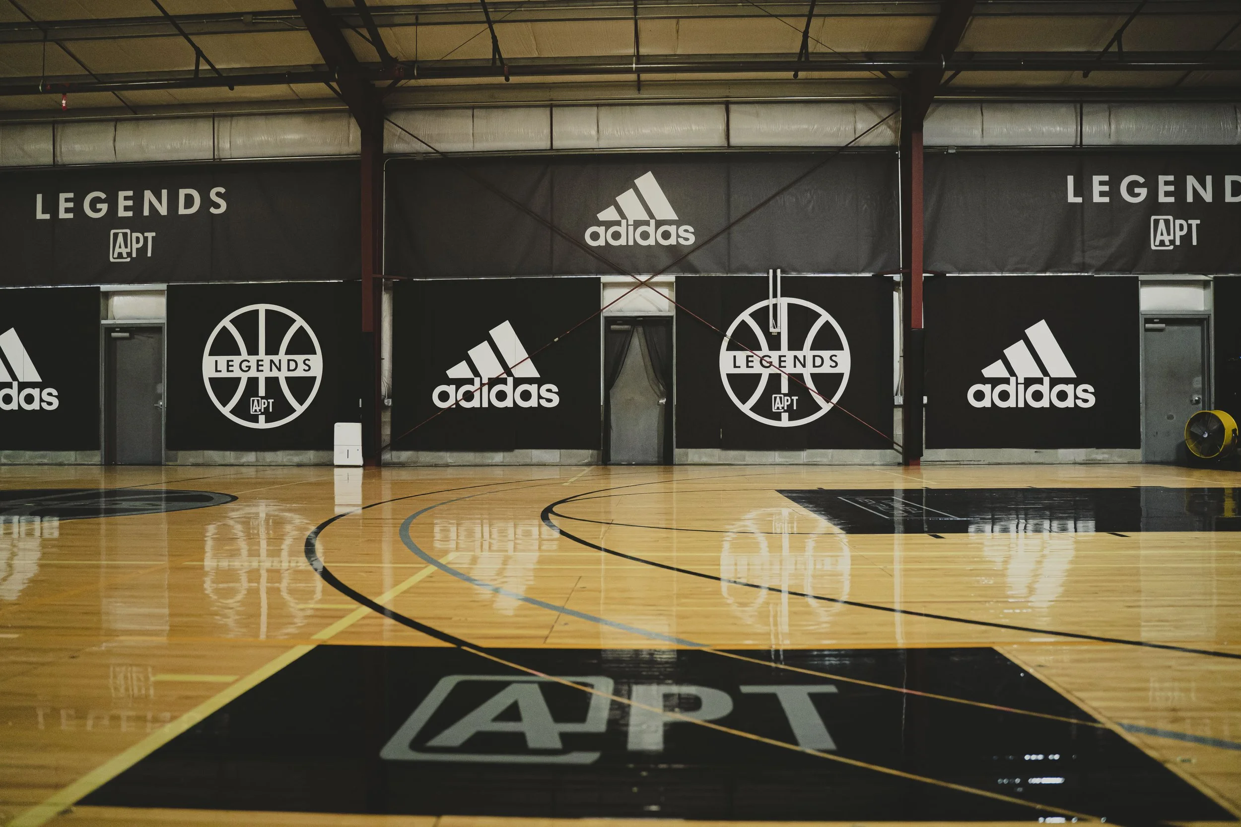 An indoor basketball court with branded banners and logos, including Adidas and Legends, along the wall. The court has distinct black, white, and yellow markings, and reflections can be seen on the polished wooden floor.