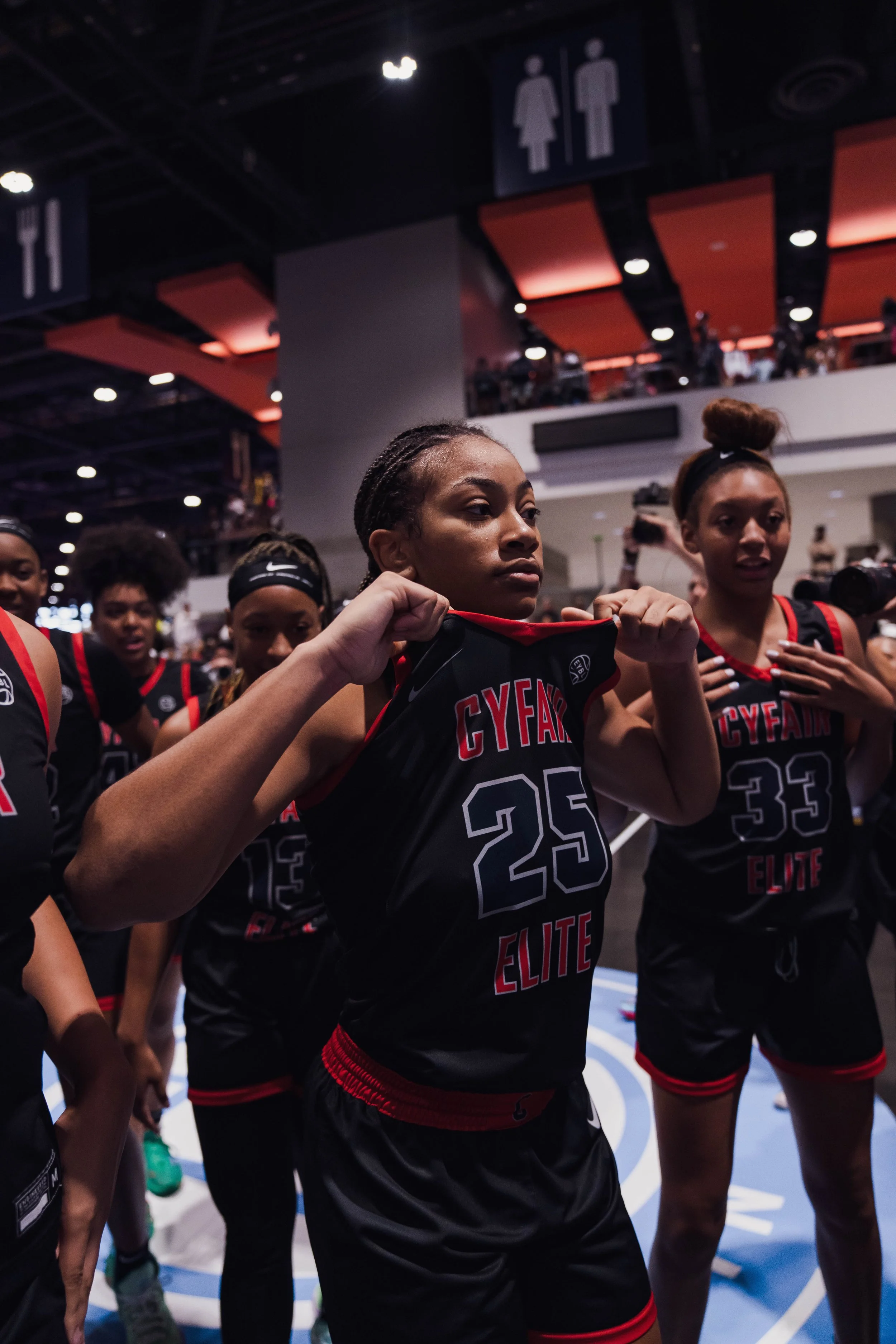 Female basketball player adjusting her jersey in locker room, surrounded by teammates, with ceiling signs indicating restrooms in background.
