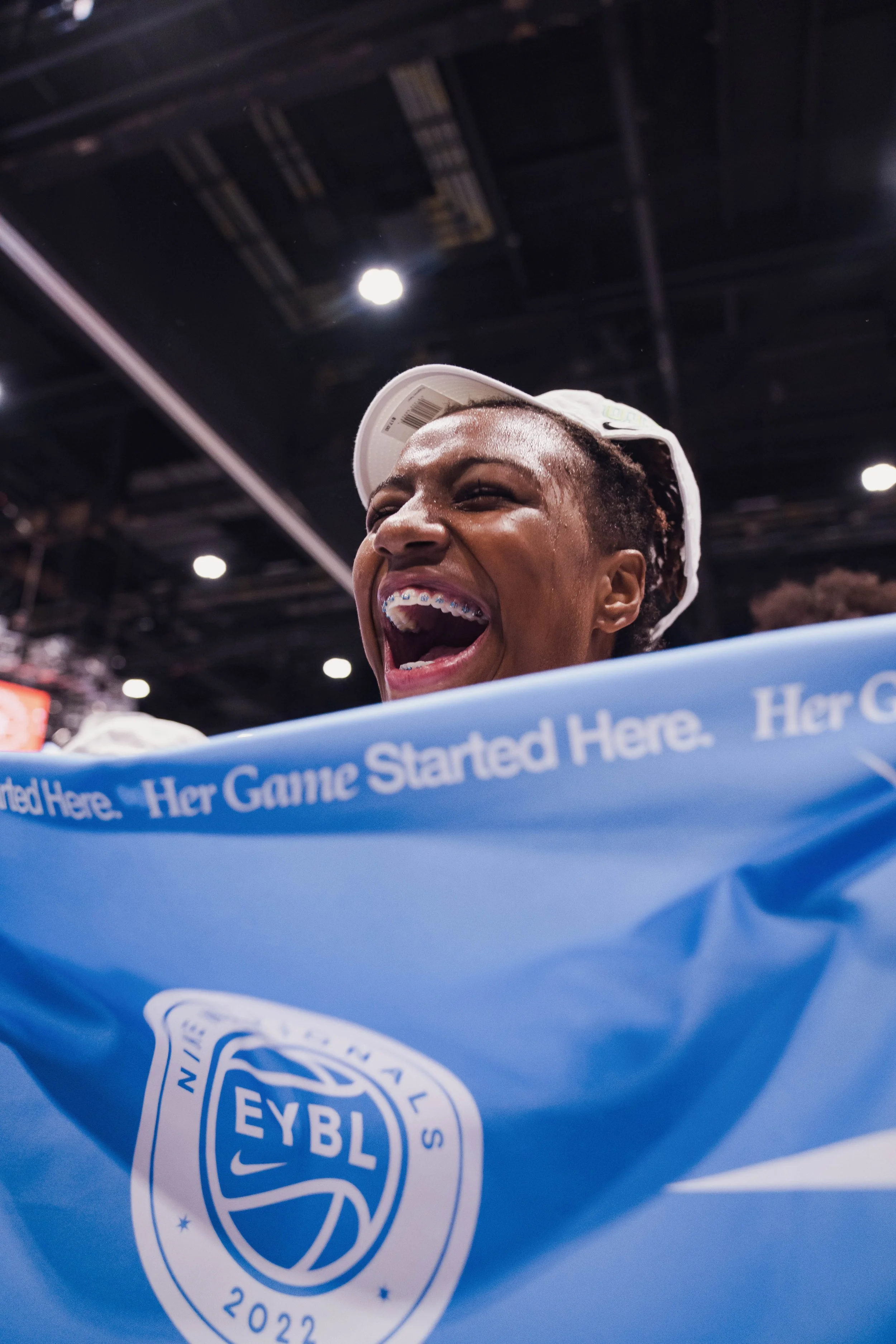 Close-up of a person's face as they laugh joyfully, wearing braces and a white cap, in a setting with overhead lights and a blue event banner with the EYBL logo. Nike.
