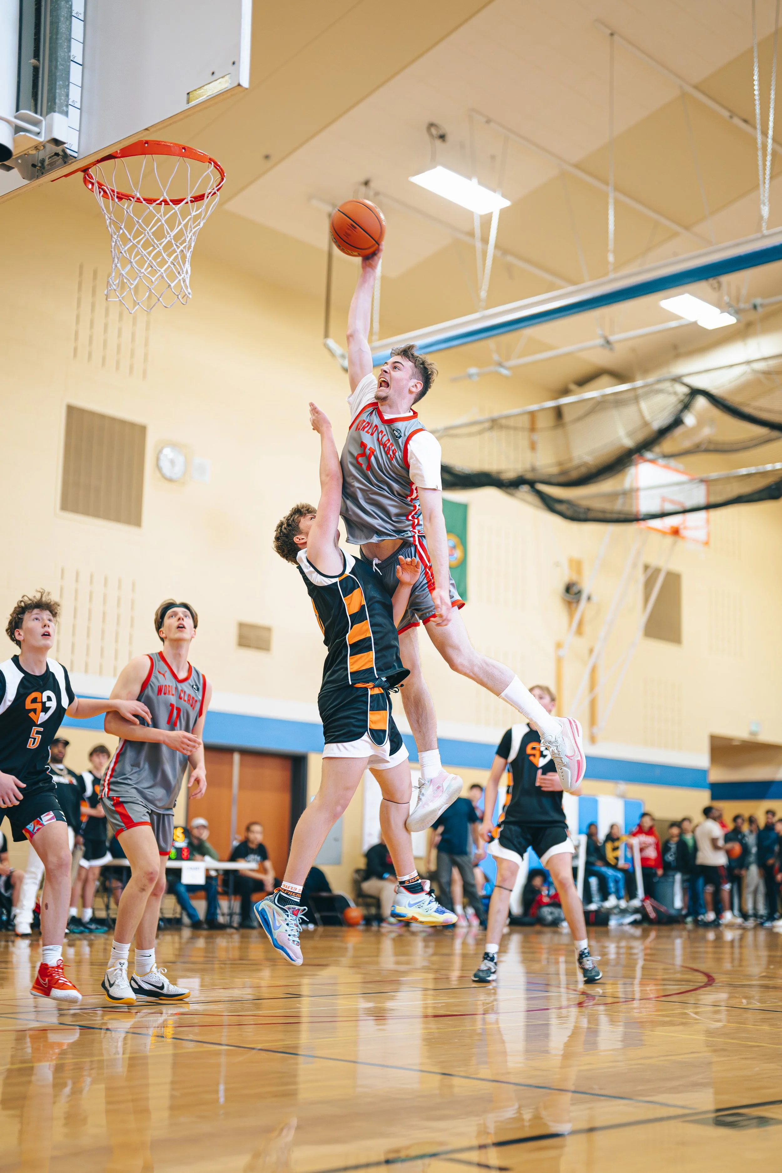 A basketball player in a jersey numbered 21 jumps to shoot the ball while an opponent attempts to block him during a game in an indoor gymnasium. Other players and spectators observe in the background.