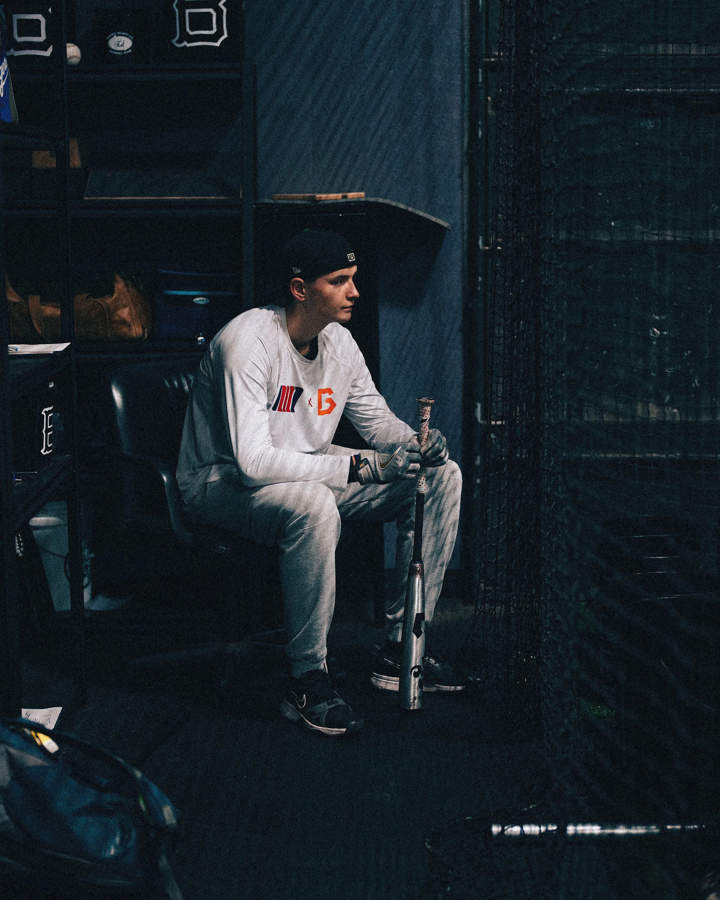 A young man wearing a white basketball jersey and a black cap sits in a dark locker room, holding a hockey stick with a patterned grip, next to a black cage.