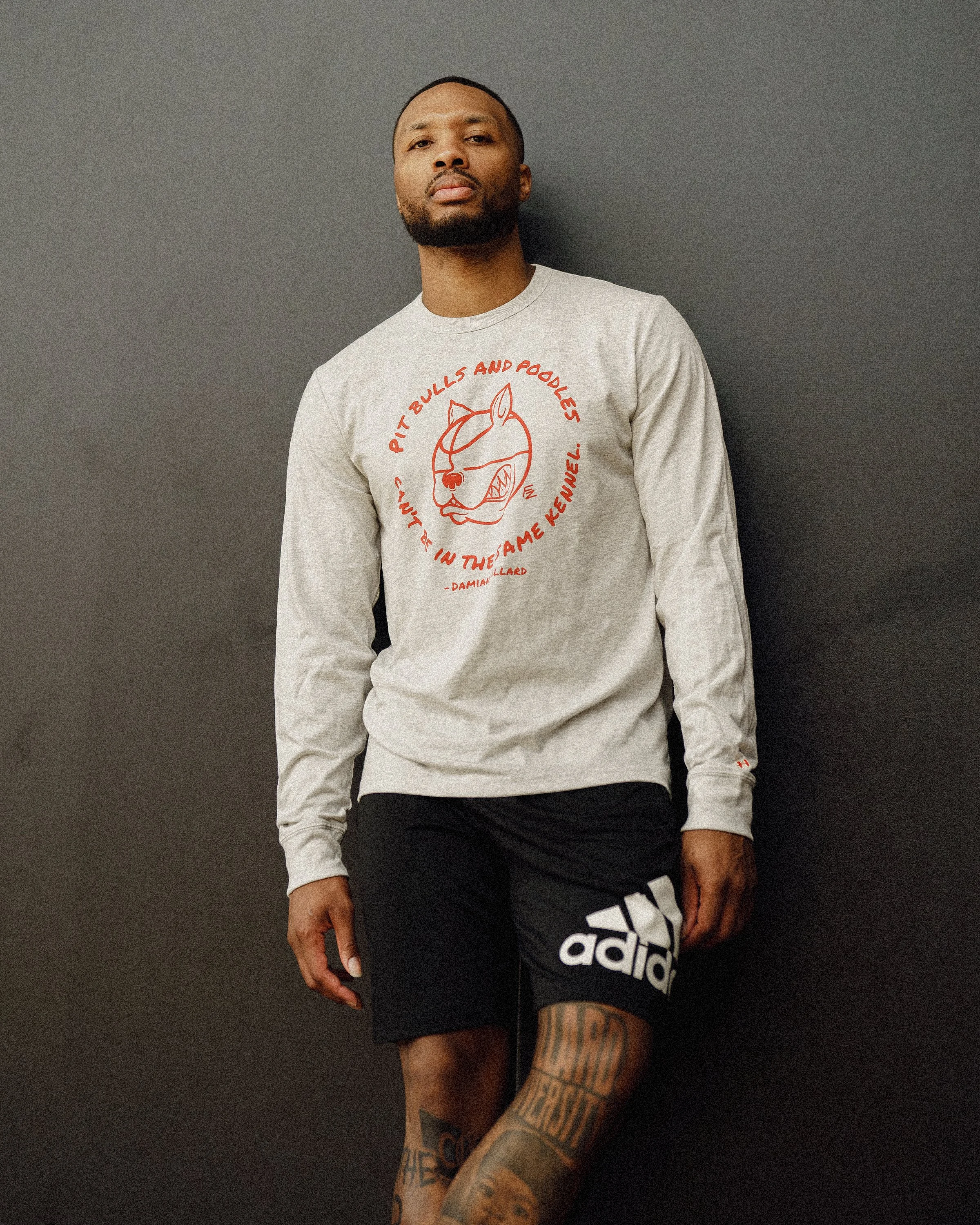 A man posing in front of a dark wall, wearing a gray sweatshirt with a red graphic and text, black shorts, and showing tattoos on his legs and arms.