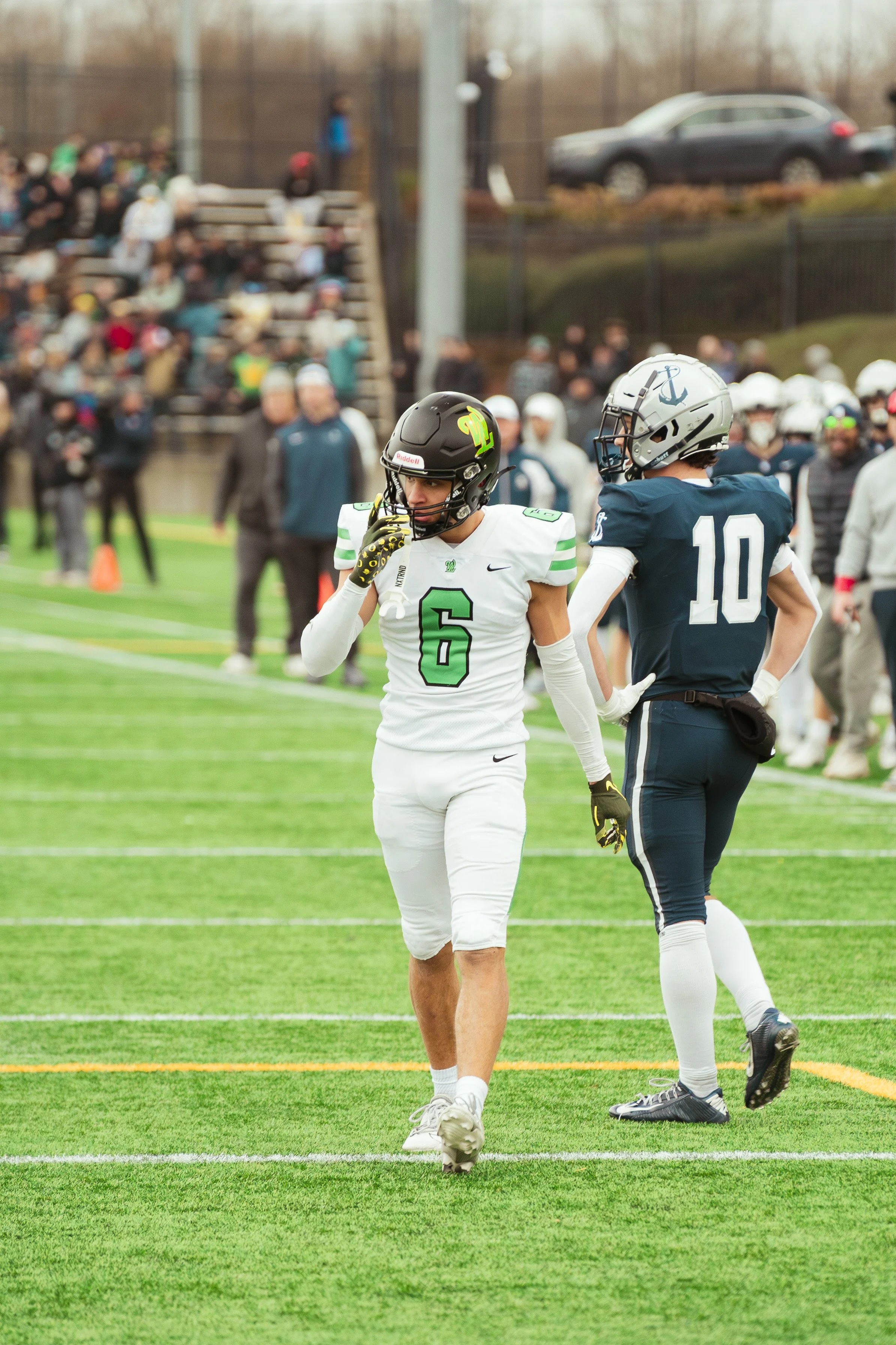 Two football players standing on the field during a game. One player is wearing a white jersey with the number 6 and a black helmet, while the other is wearing a navy jersey with the number 10 and a silver helmet with an anchor logo. There are specta