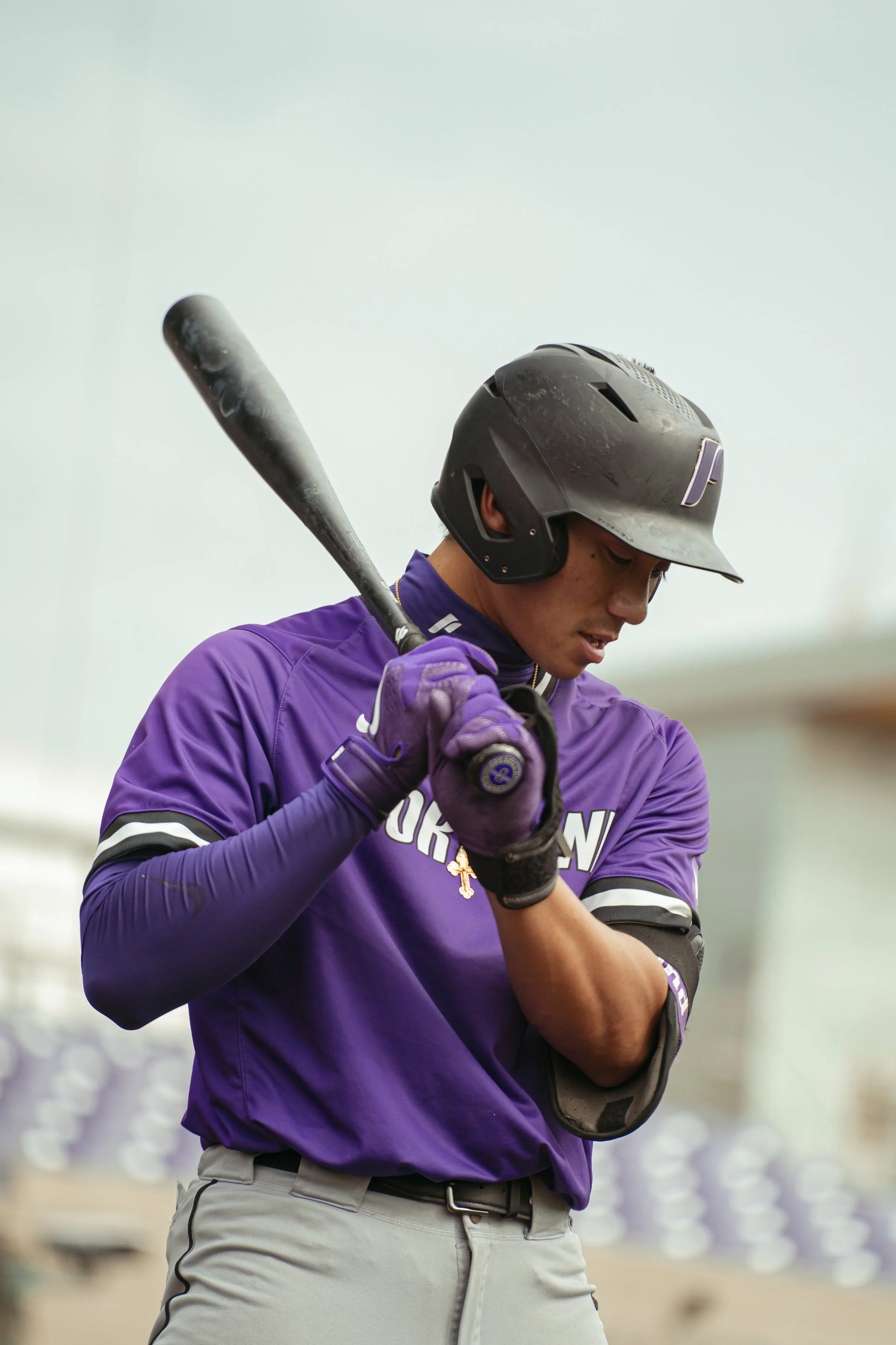 A person in a purple sports jersey, gray baseball pants, and a black batting helmet holding a baseball bat, preparing to bat in a baseball game.