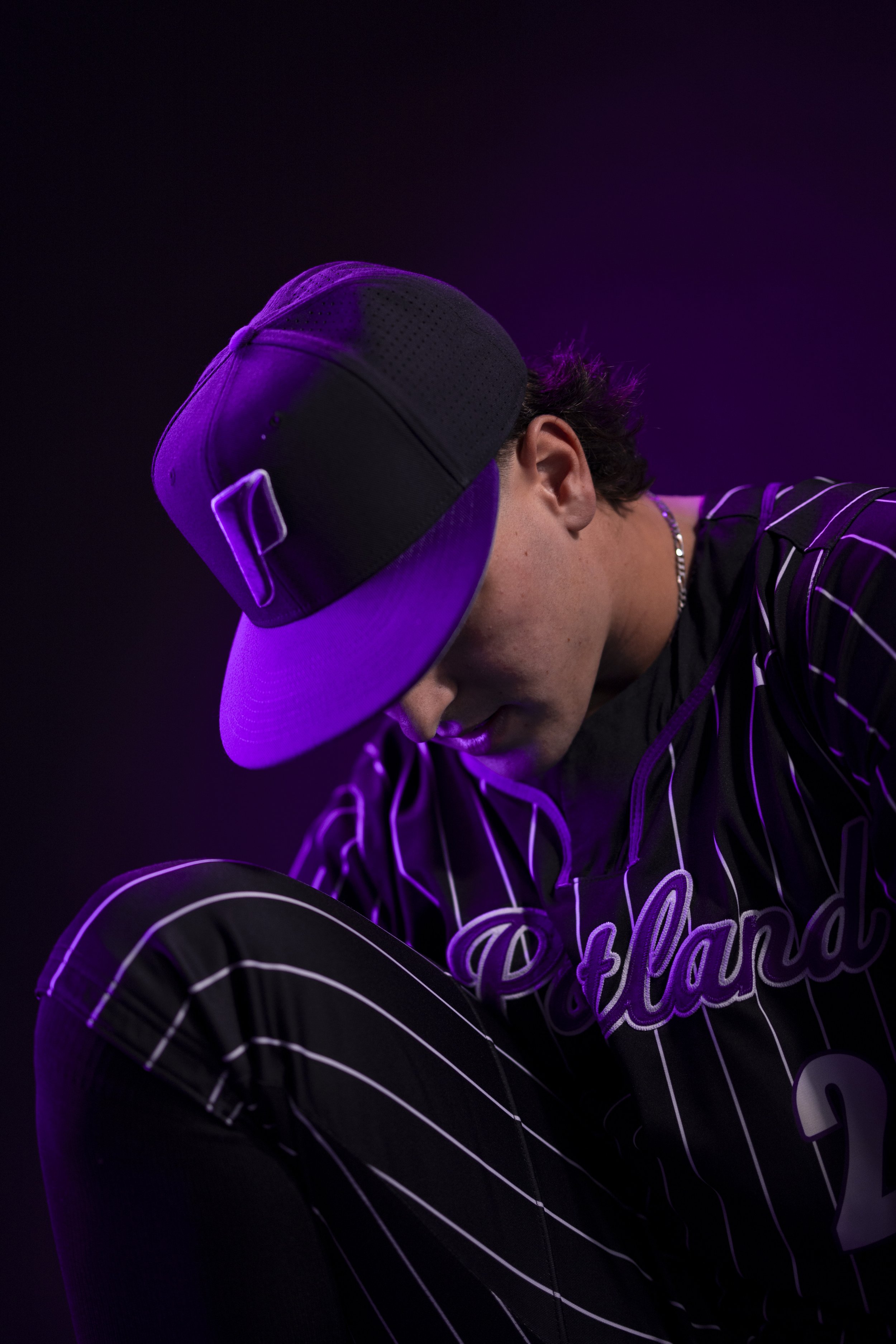 A young man wearing a gray baseball cap, black and purple striped shirt, and black pants with white stripes, sitting against a dark background illuminated by purple lighting.