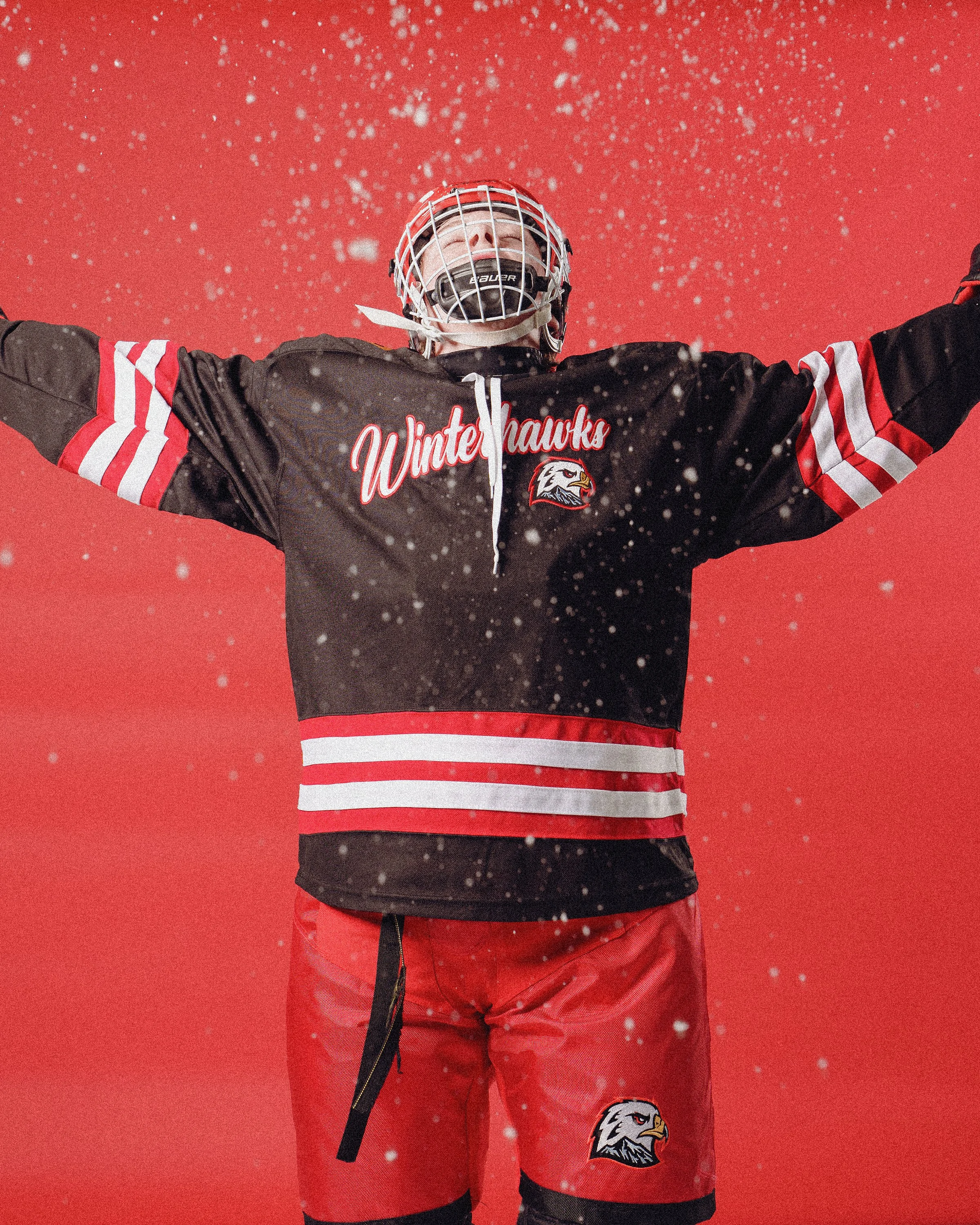 A person wearing a black and red hockey jersey with the word "Winterhawks" and a hawk logo, red hockey pants, a white helmet with a face cage, and holding a hockey stick, standing against a red background with snow falling.