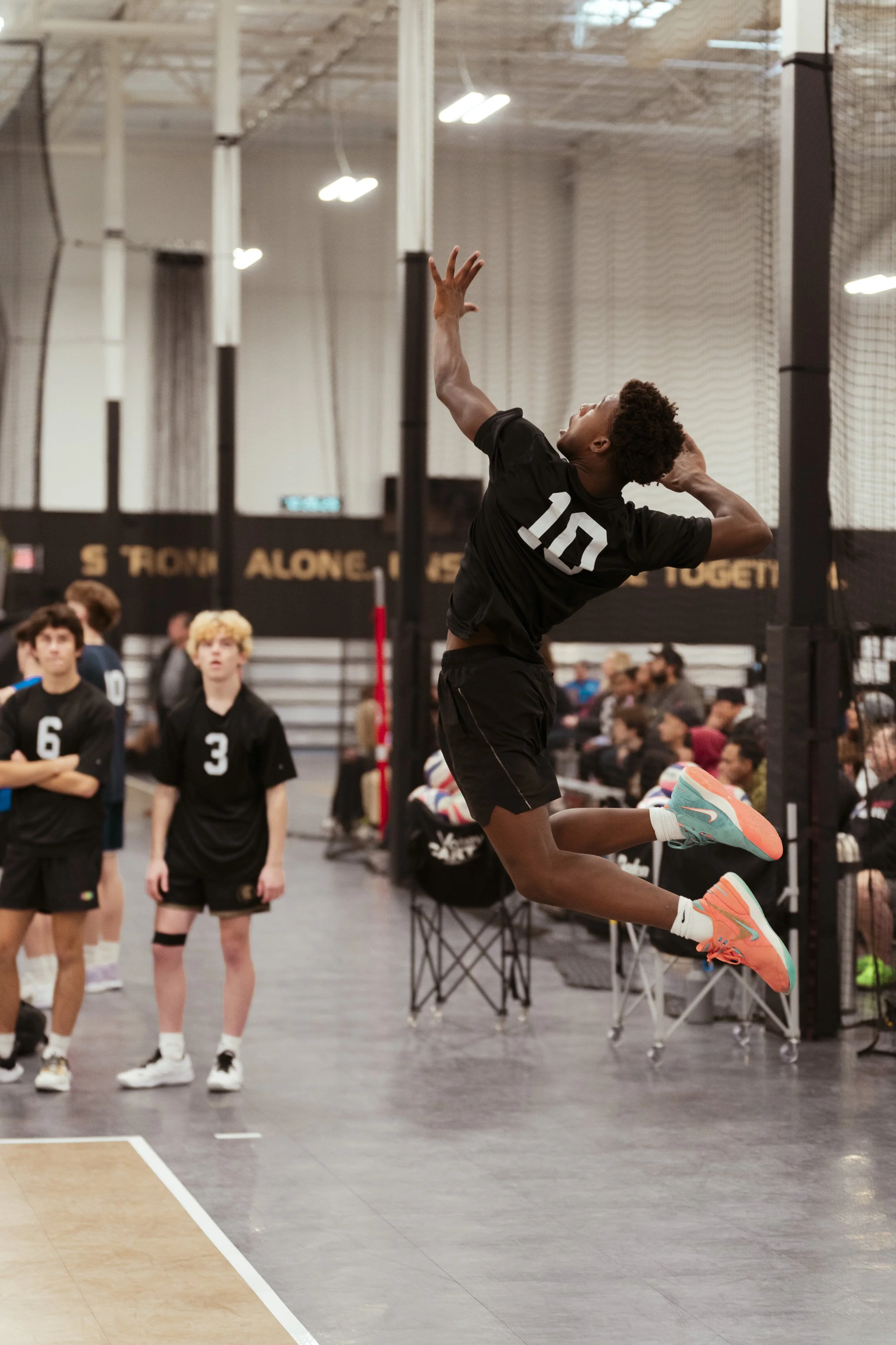 A male volleyball player in mid-air jumping to spike the ball during a game in an indoor gym with spectators and teammates watching.