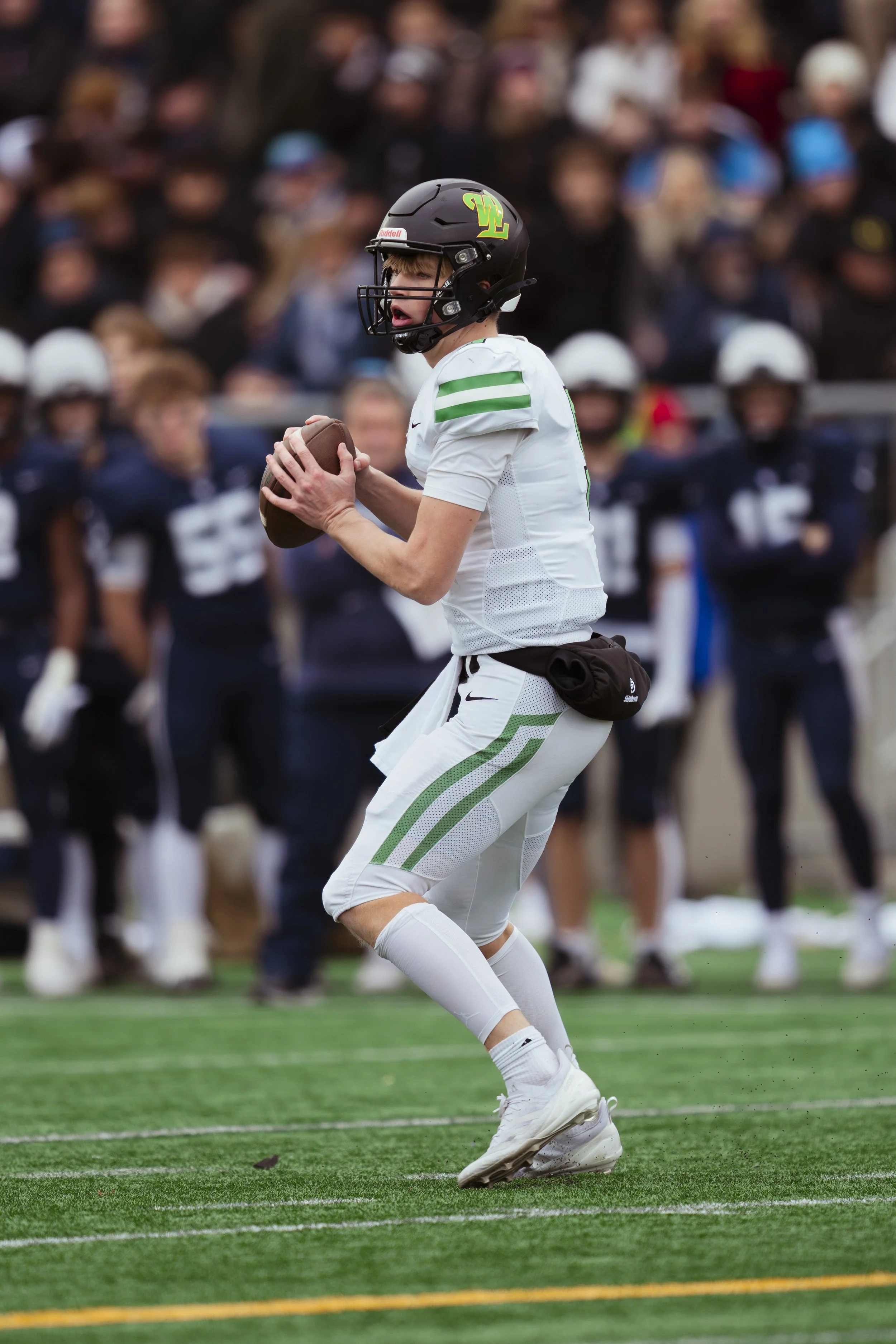 A young football player in a white uniform with green stripes, wearing a black helmet, catches a football on a green field with yellow and white lines, with a crowd of spectators in the background.