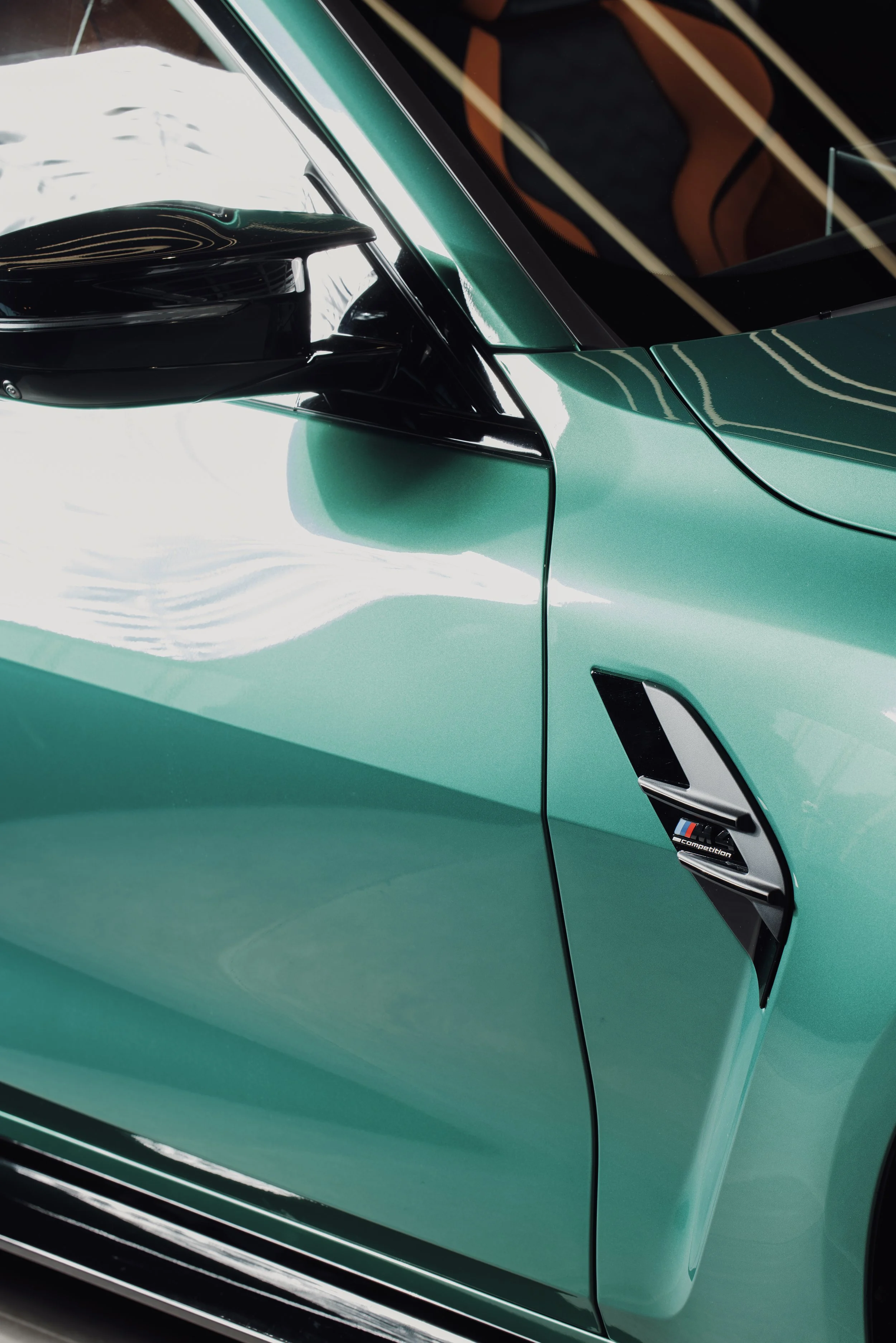 Close-up of a teal-colored luxury sports car's side, featuring a black side mirror, a vent with a BMW M logo, and reflections of ceiling lights.