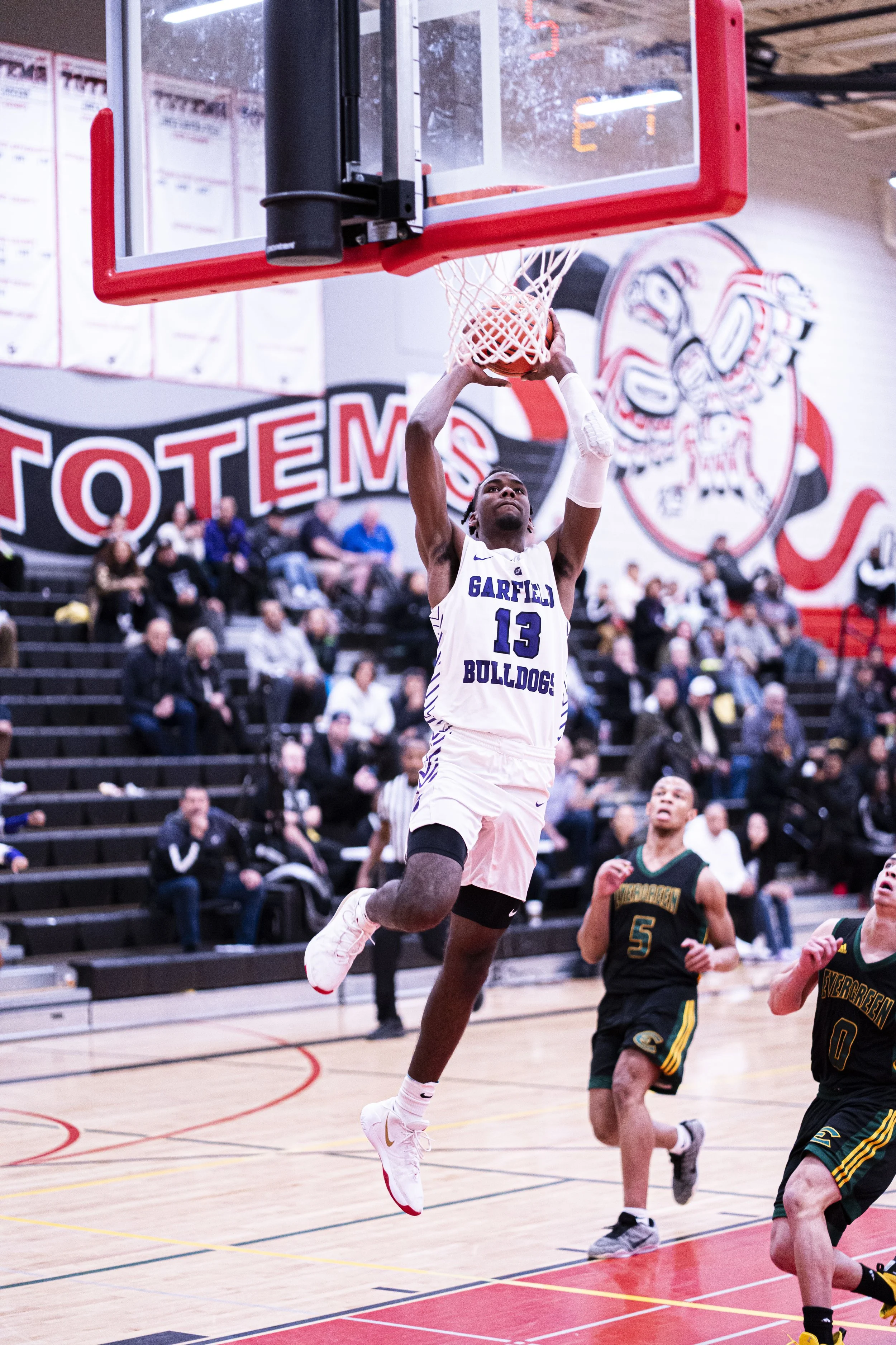 A basketball player wearing a Garfield Bulldogs jersey with the number 13 is jumping towards the basket for a dunk in an indoor gym, with spectators watching from the bleachers.