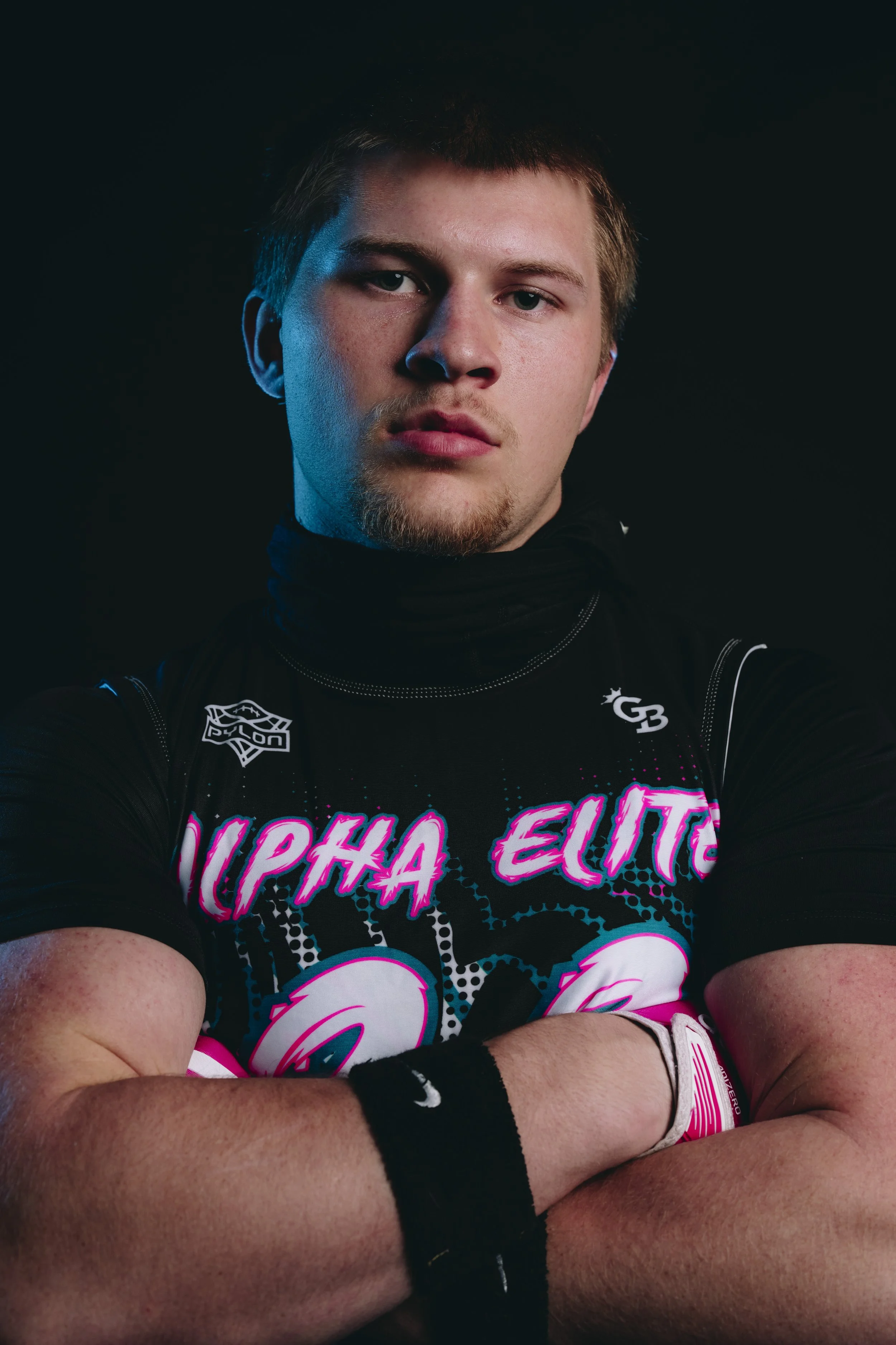 A young man with short light brown hair and a light beard is wearing a black sports jersey with pink and white lettering, black wristbands, and a pink wristband on his left wrist. He has a serious expression and is crossing his arms in front of him, 