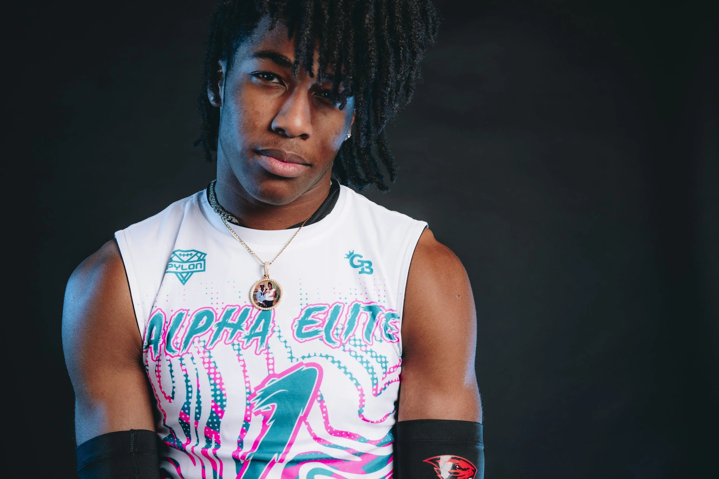Young female athlete with natural hair wearing a white sports jersey with pink and blue graphics, a gold necklace with a pendant, black arm sleeves, and standing against a dark background.