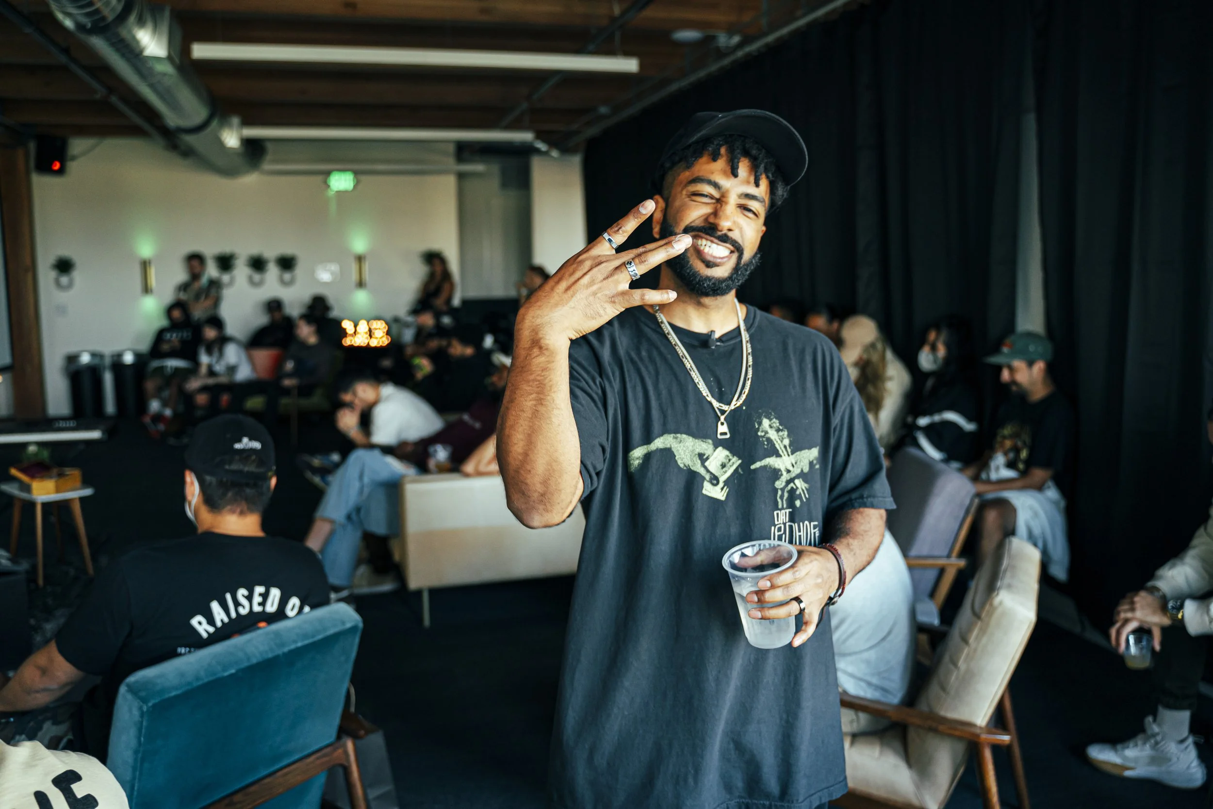Smiling man with black beard and mustache, wearing a black cap, black t-shirt, and layered necklaces, holding a plastic cup, making a peace sign with his right hand in a group setting with people sitting and standing in a casual indoor event.