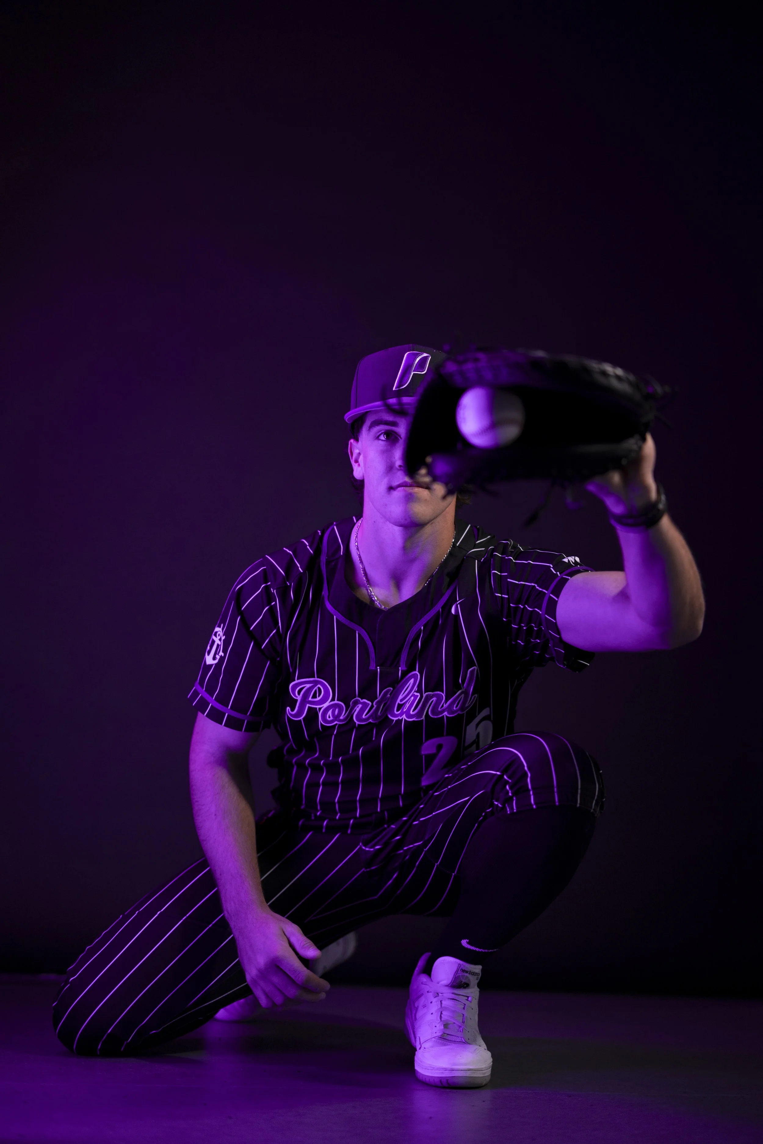 A young man in a Washington Nationals baseball uniform is kneeling on the floor and holding a baseball glove with a baseball inside, against a dark background with purple lighting.