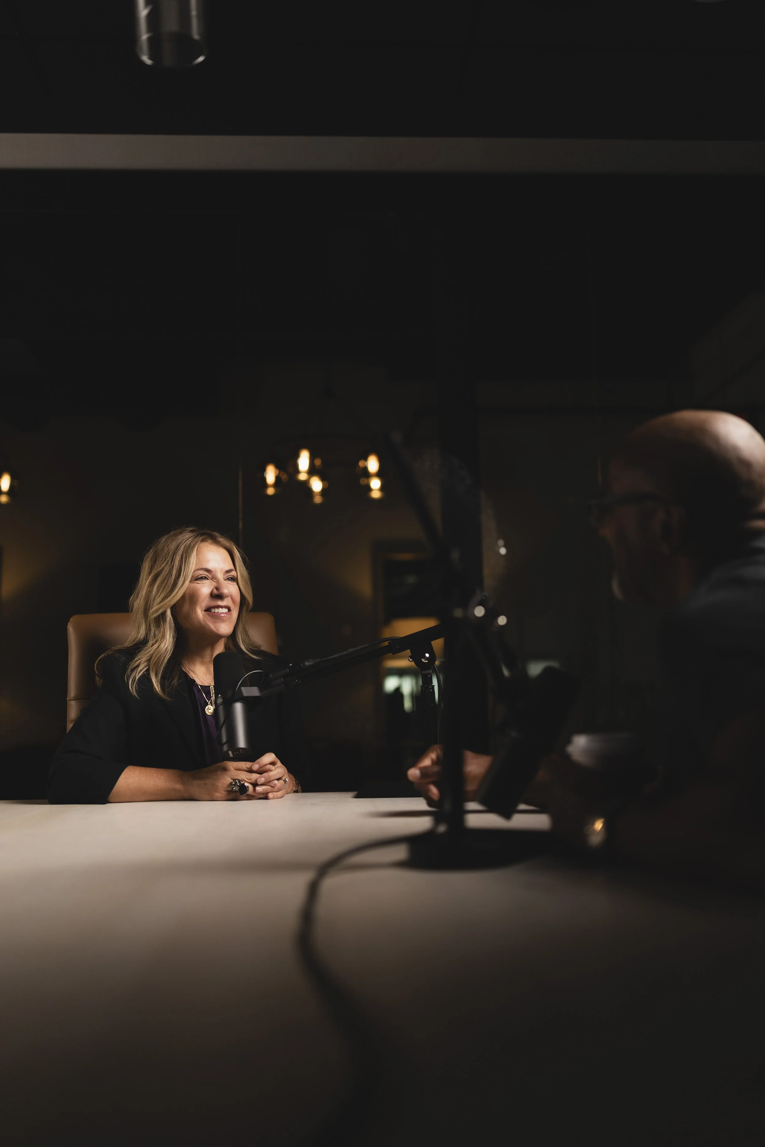 Woman with blonde hair smiling, seated at a table, speaking into a microphone during an interview or podcast, with an older man with glasses faintly visible on the right, against a dim background with warm lighting.
