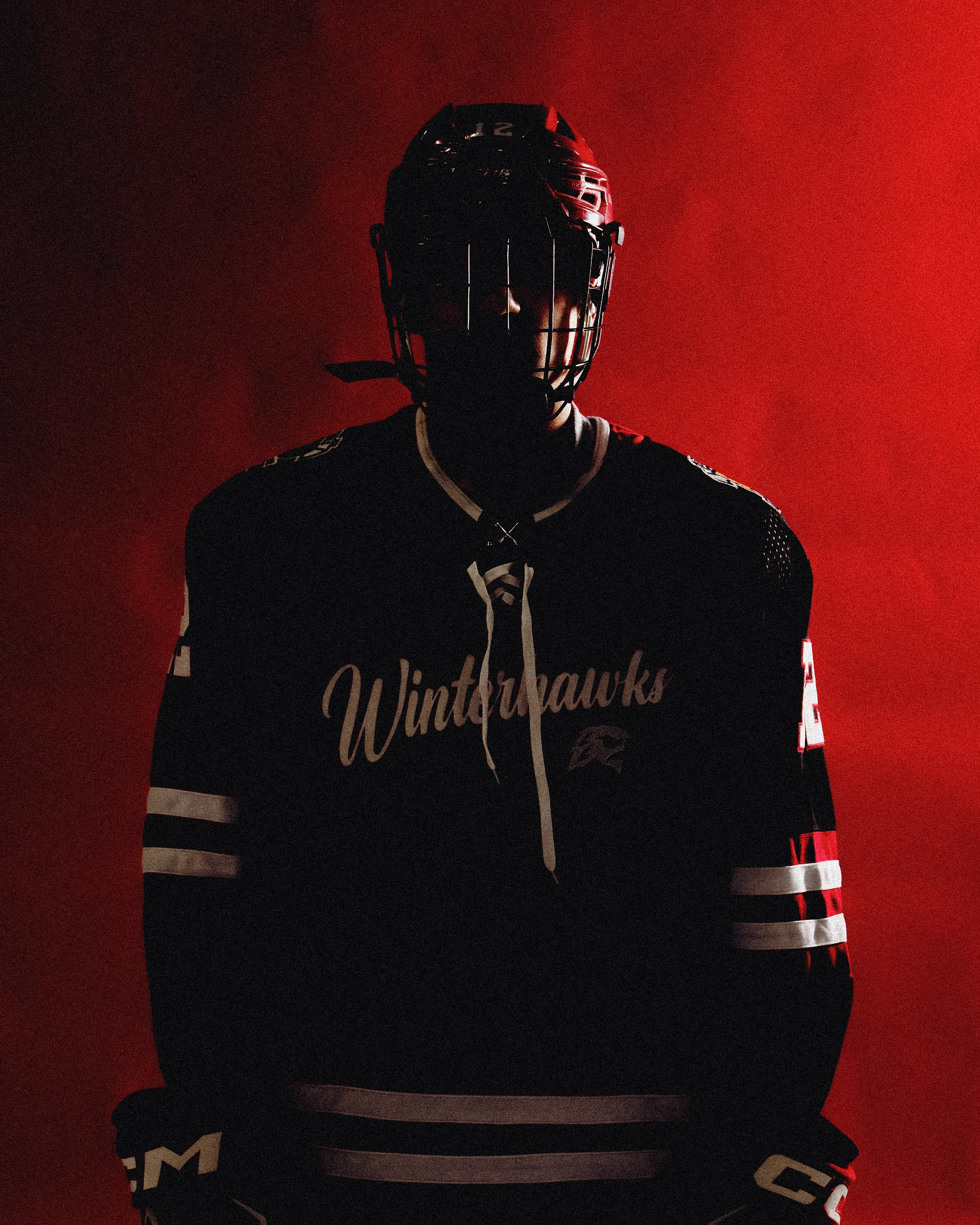 Hockey player wearing a black jersey with white and red accents, a black helmet with a cage, standing against a red background.