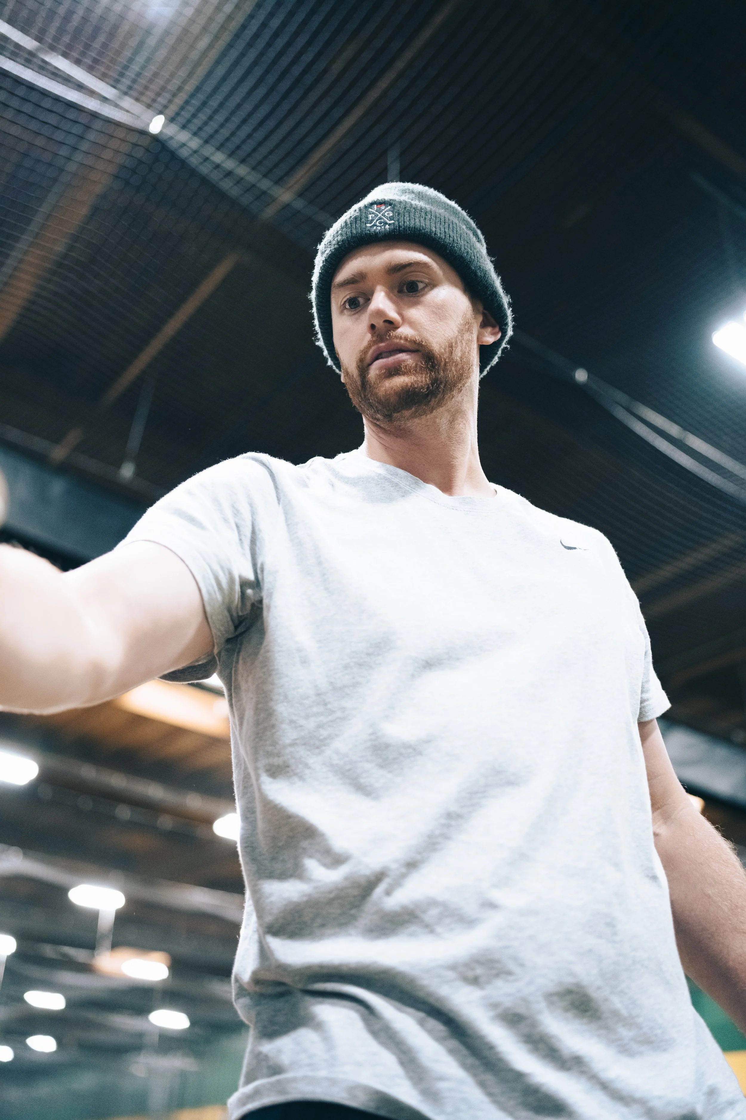 A man with a beard wearing a gray beanie and a light gray t-shirt in an indoor gym or sports facility.
