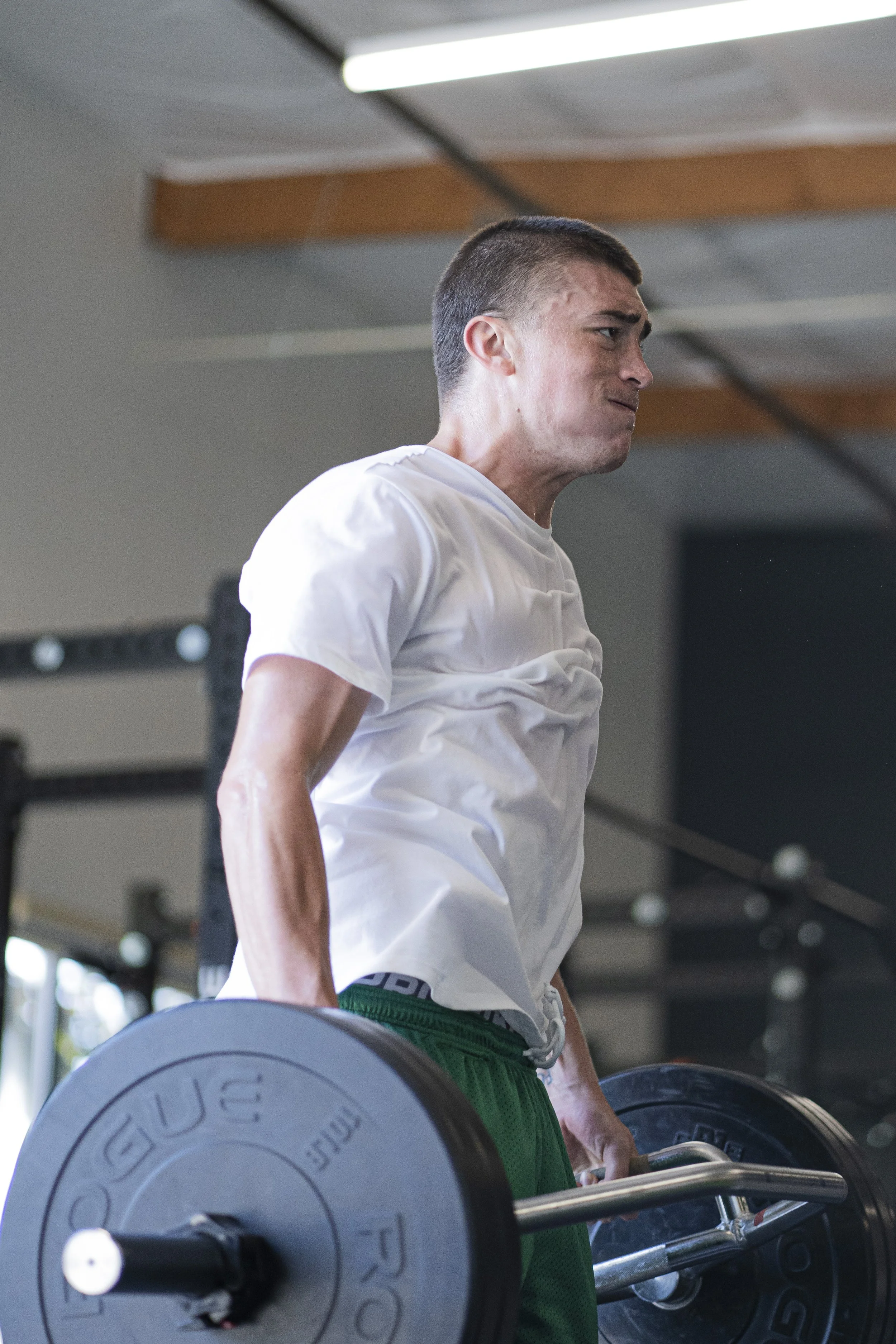 A man with short hair and a white shirt is lifting a barbell with weights at a gym. He is focused and looking slightly upward. The gym has a ceiling with wooden paneling and fluorescent lights.