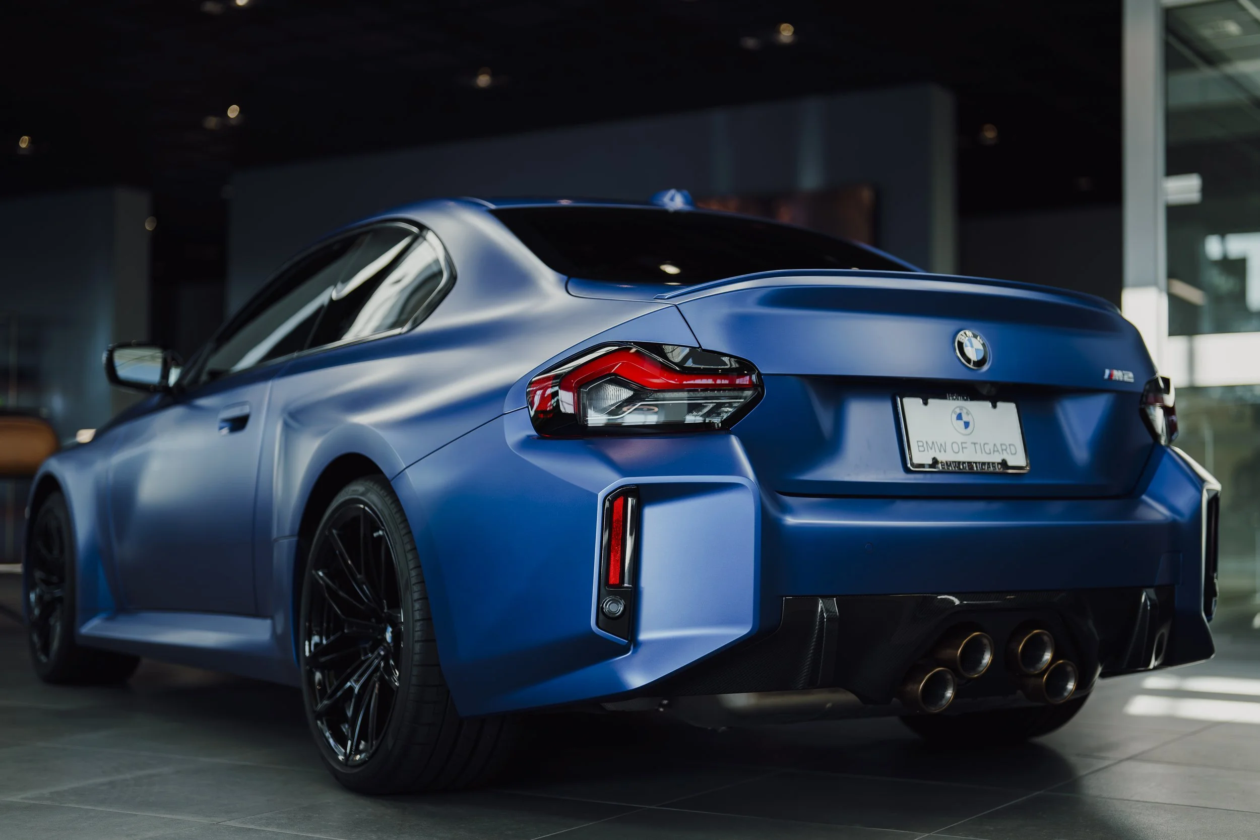 A blue BMW sports car displayed indoors in a showroom, showing the rear and side view with sporty design, black wheels, dual exhaust pipes, and a license plate that reads 'BMW OF TIGARD'.
