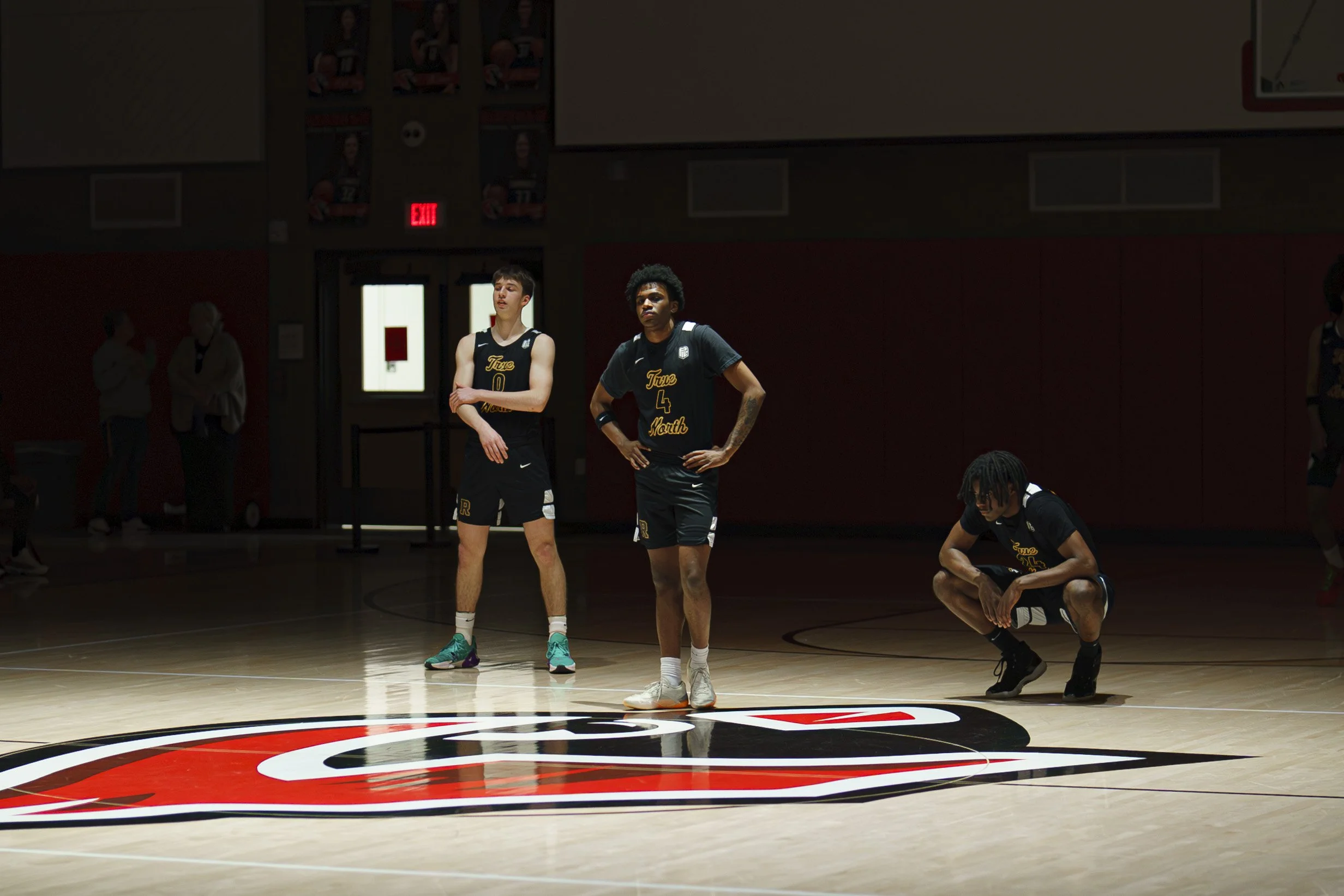 Three basketball players, two males and one female, standing on a basketball court with a red and black logo, wearing black jerseys with yellow text, appear to be reflecting during a game or practice. There are two other people and banners in the bac
