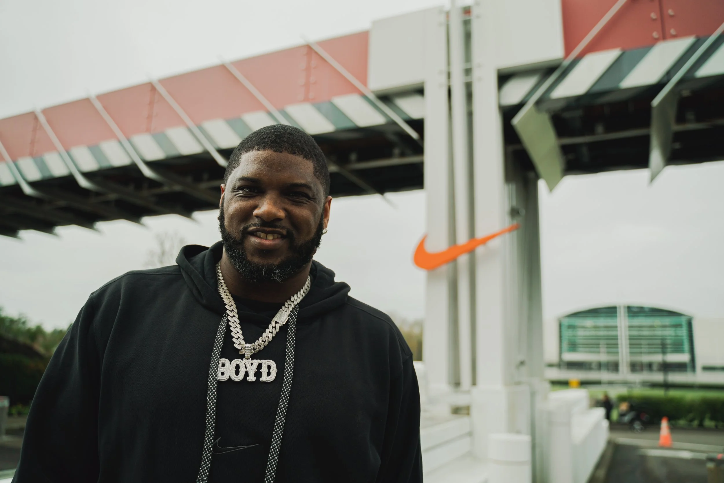 A young man with a beard and short hair smiling outdoor near a modern building, wearing a black hoodie and large silver chain with pendant.