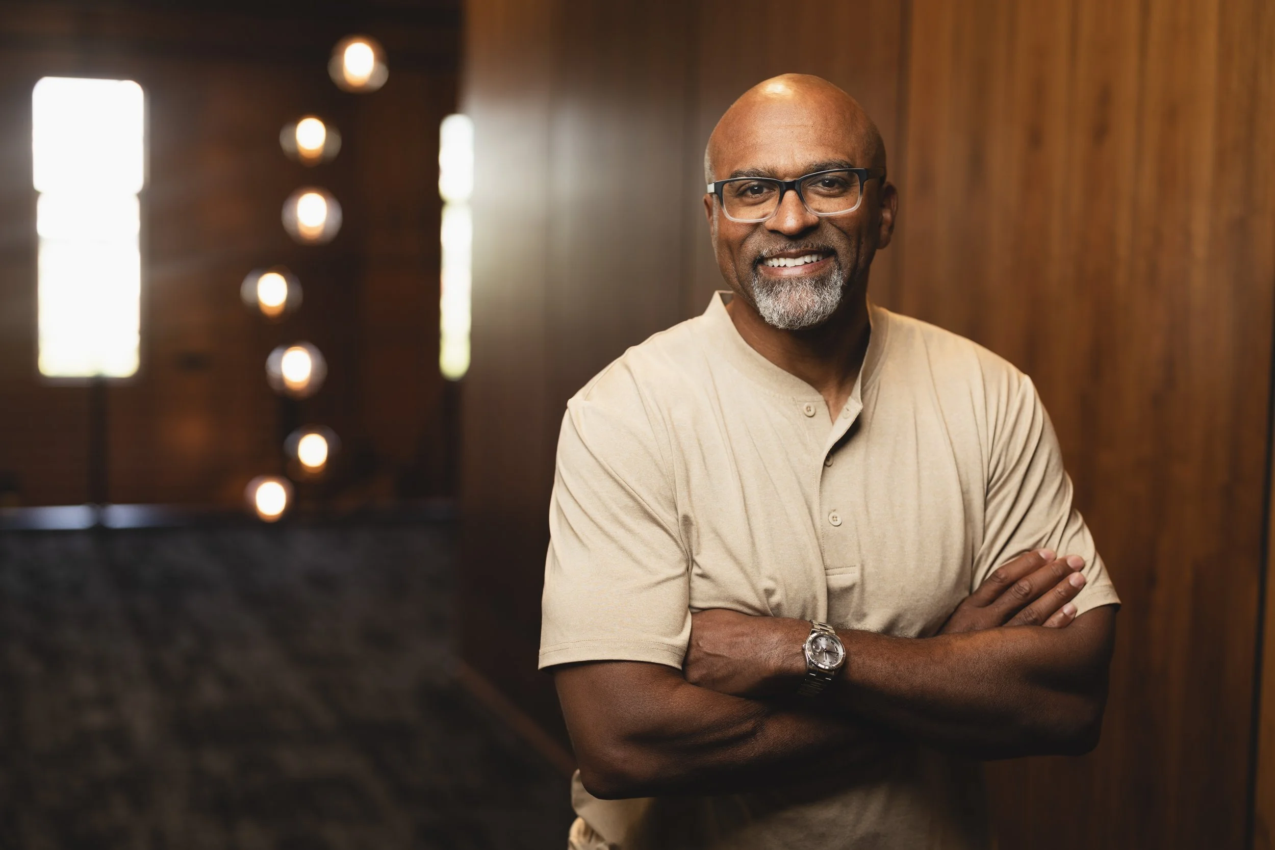 A smiling middle-aged African American man wearing glasses and a beige shirt, standing with arms crossed against a wooden wall with warm lighting.