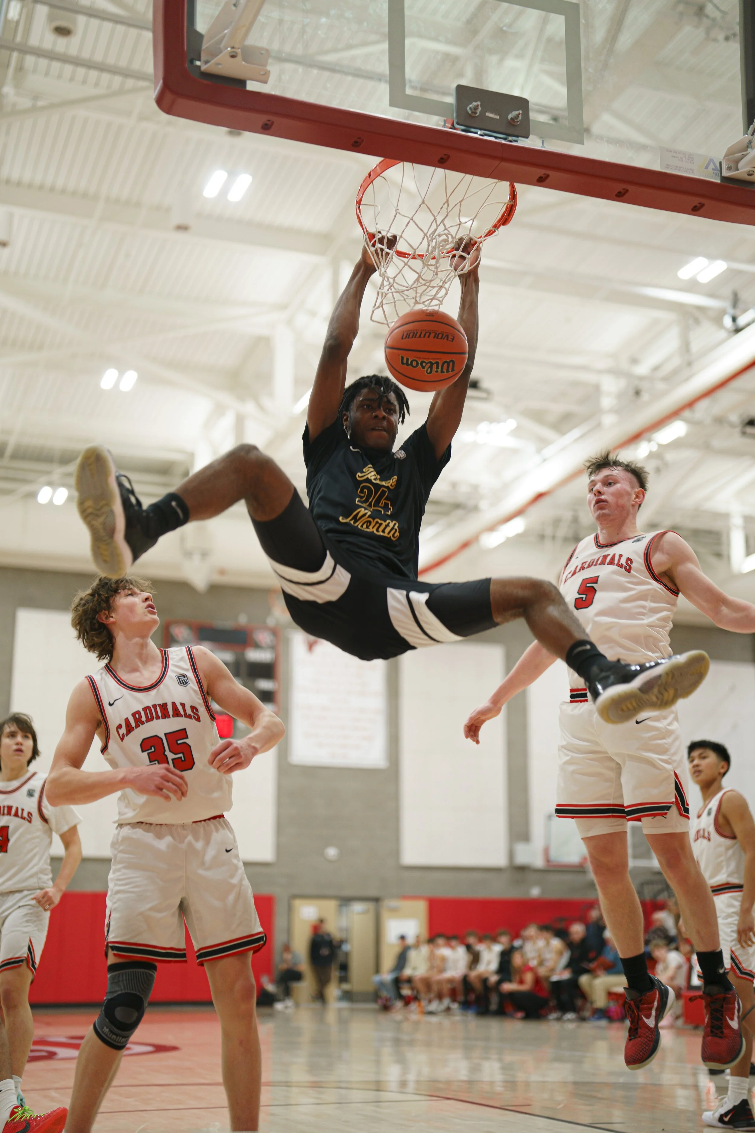 A basketball player in a dark uniform is jumping to dunk the ball into the hoop, while several players in white uniforms with red accents and the word 'Cardinals' are nearby on the court in a gymnasium.
