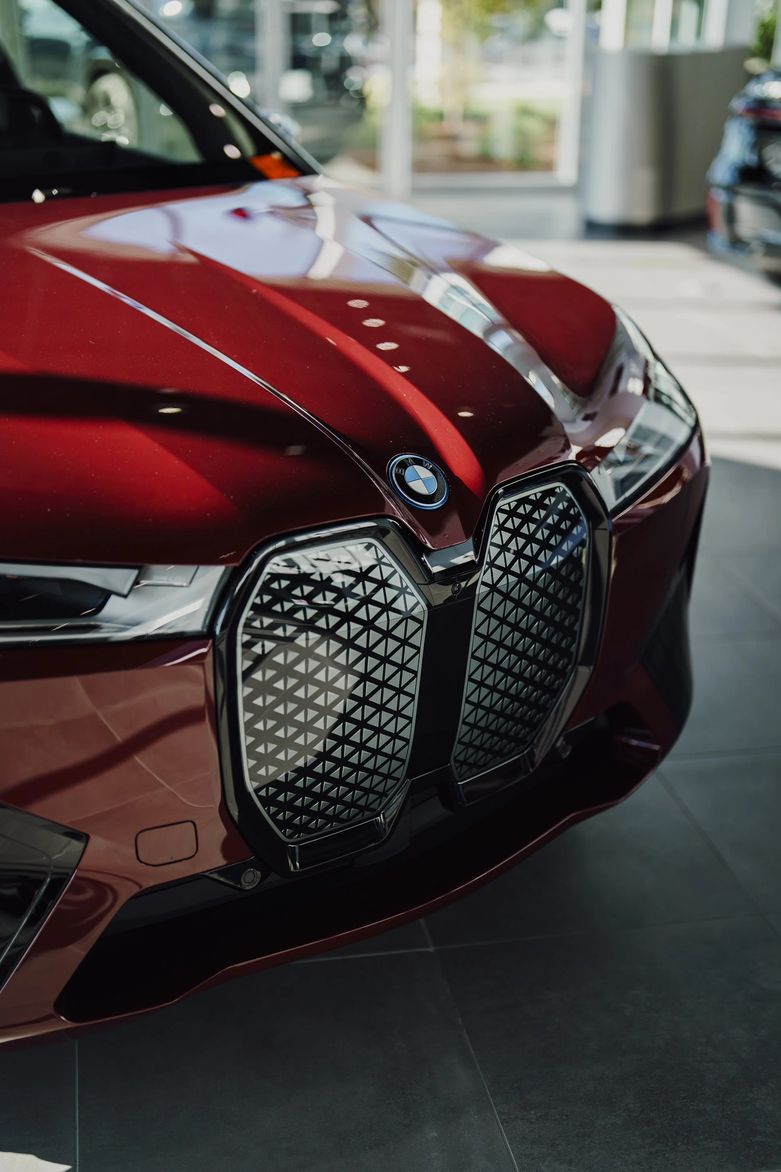 Close-up of a red BMW car with distinctive grille, inside a car dealership showroom, with reflections of ceiling lights on its hood, and other cars visible in the background.