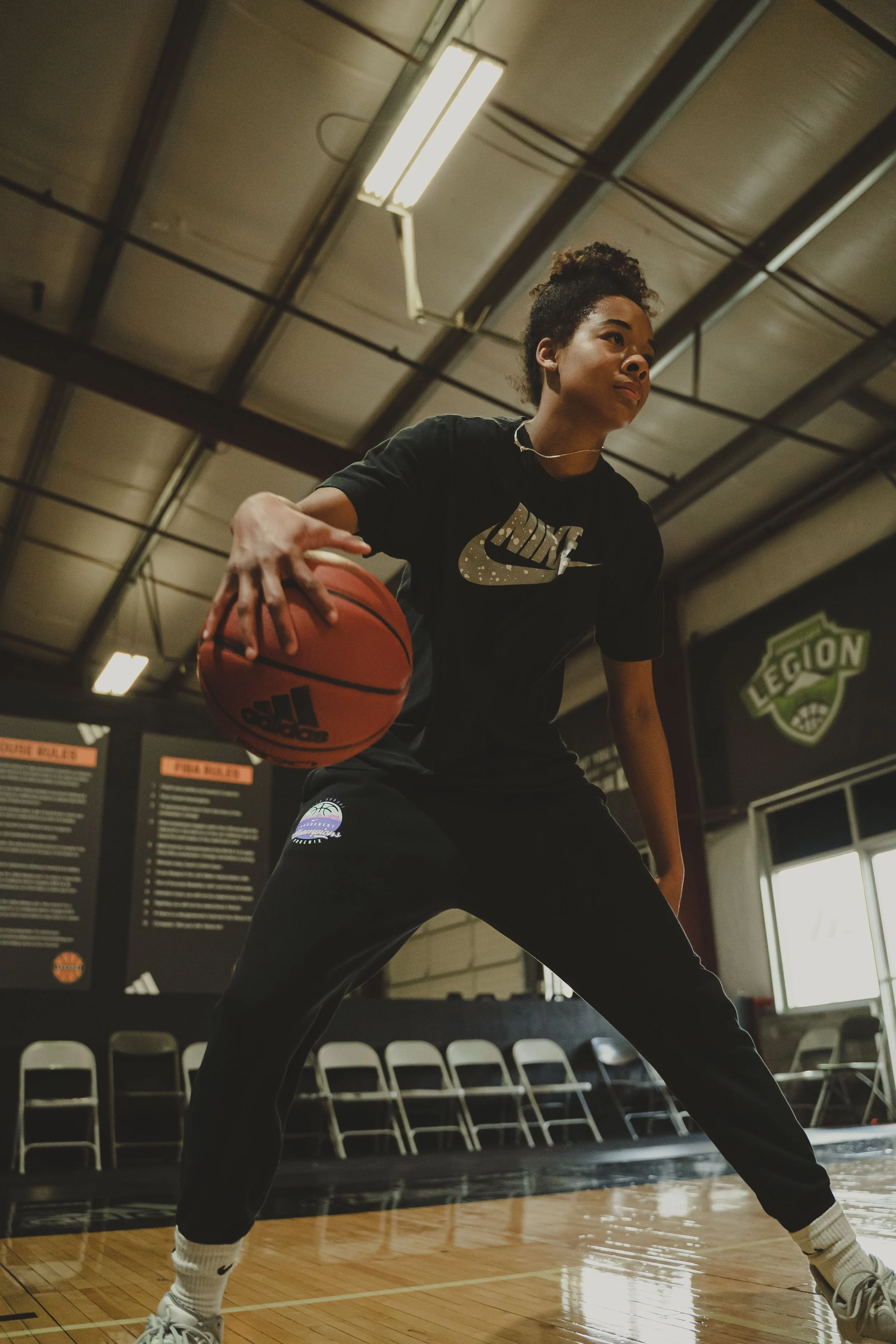 A female basketball player in a gym dancing while holding a basketball, wearing a black Nike t-shirt, black pants, white Nike socks, and white sneakers. The background shows a gym with chairs, a large window, and a wall with signs.