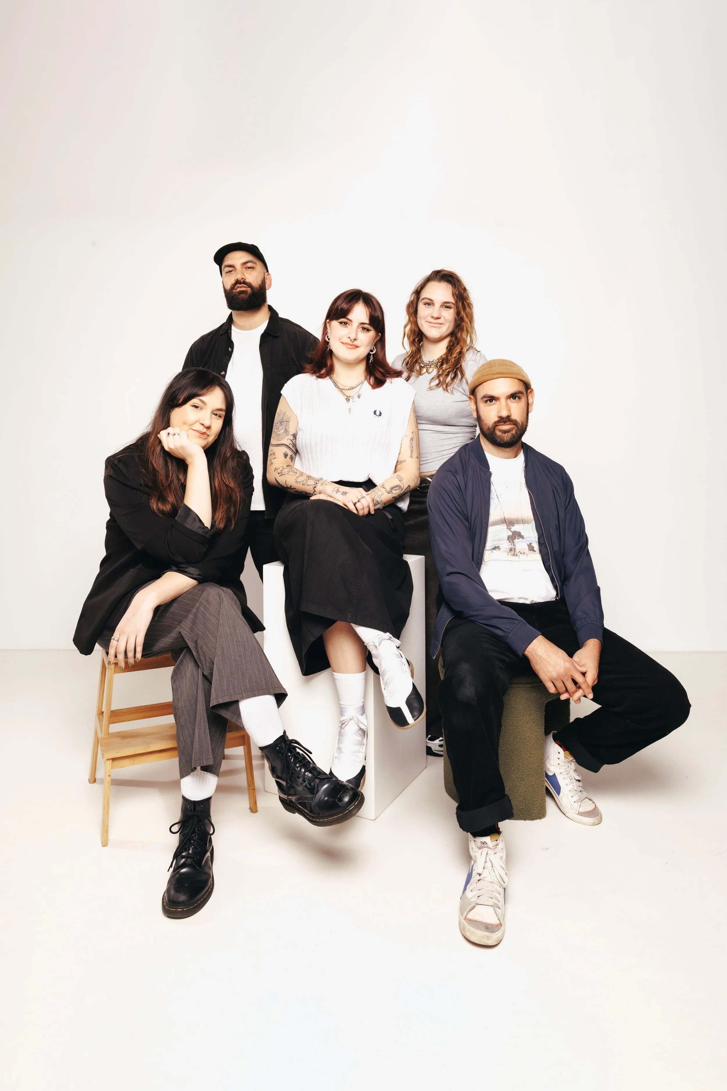 Group of five young adults; the BASE MGMT team, lead by Sinead O'Hara and Kori Reay-Mackey, posing together in a photo studio with a plain white background, sitting and standing.