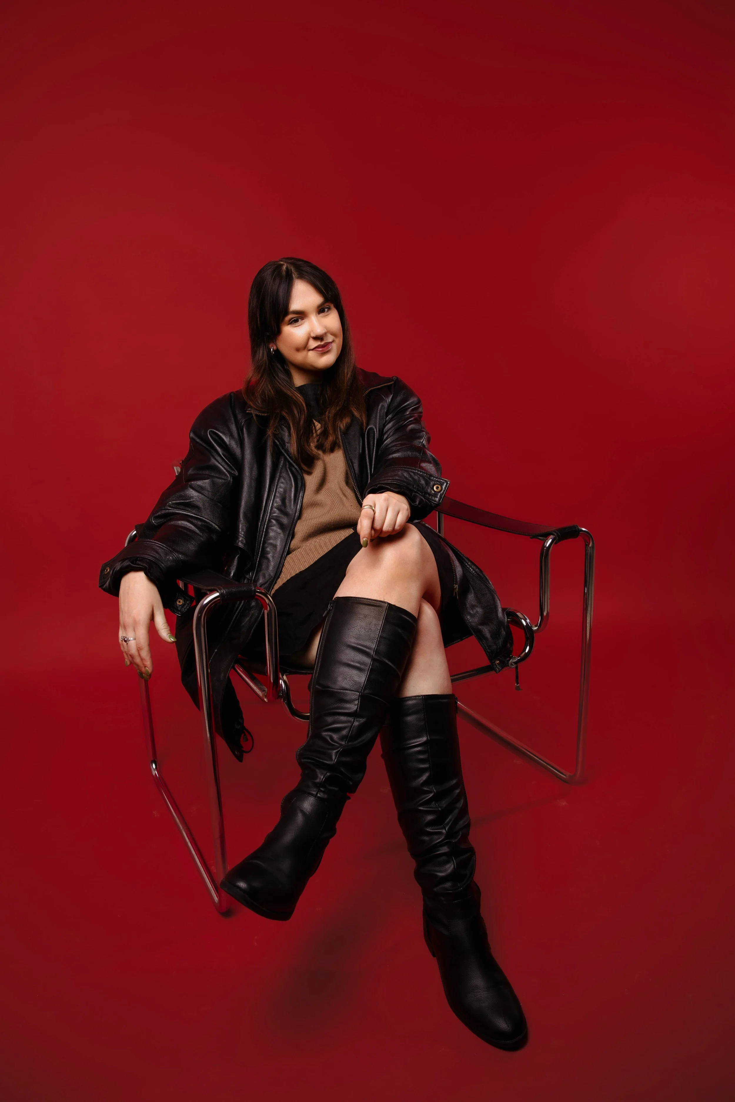 Sinead O'Hara, Founder & Artist Manager at BASE MGMT, sits on a chair against a red backdrop wearing a leather jacket and boots.