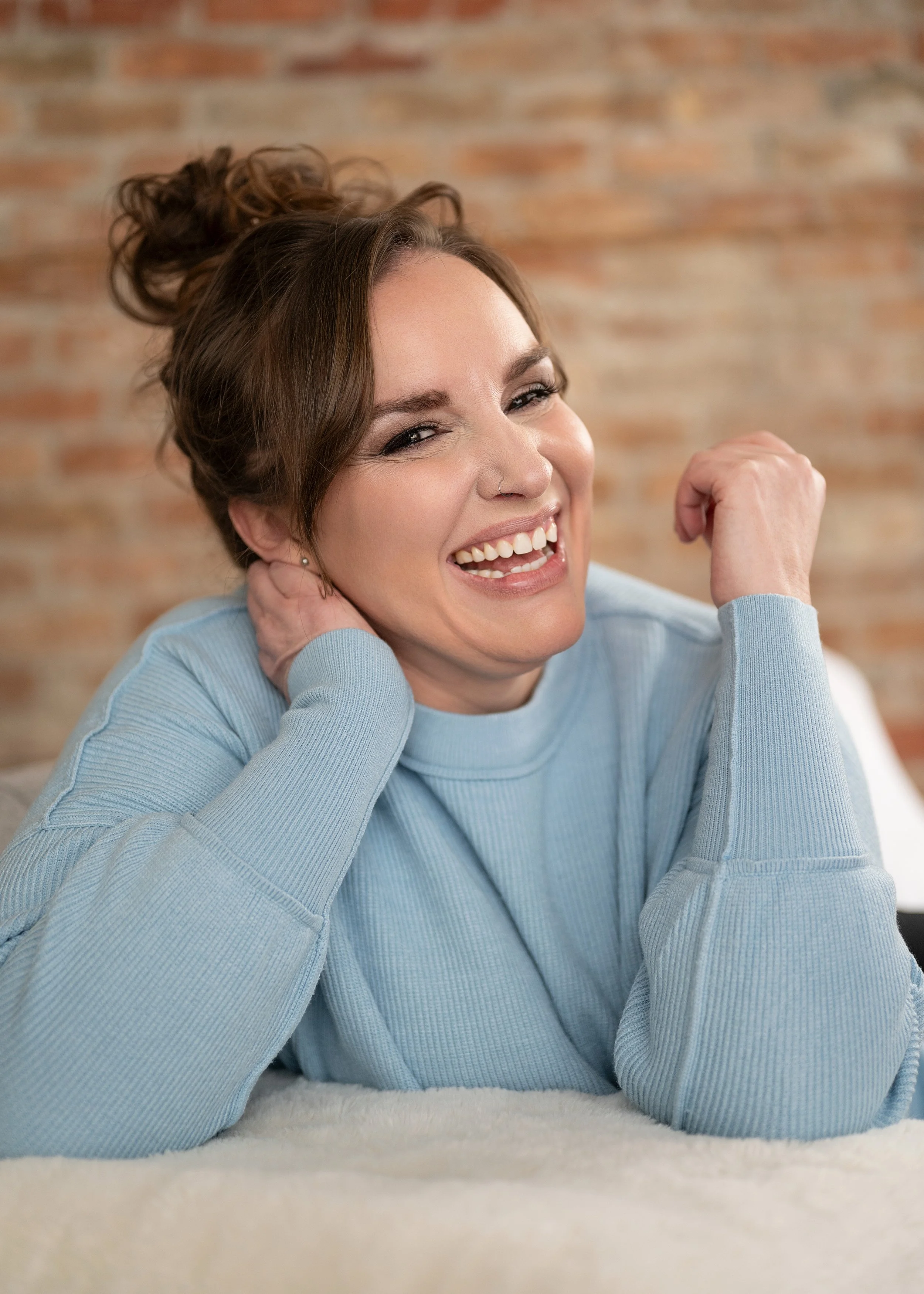 A personal brand photograph of a woman in a light blue sweater is smiling and appearing joyful, with one hand behind her head and the other resting on her neck, in front of a brick wall.