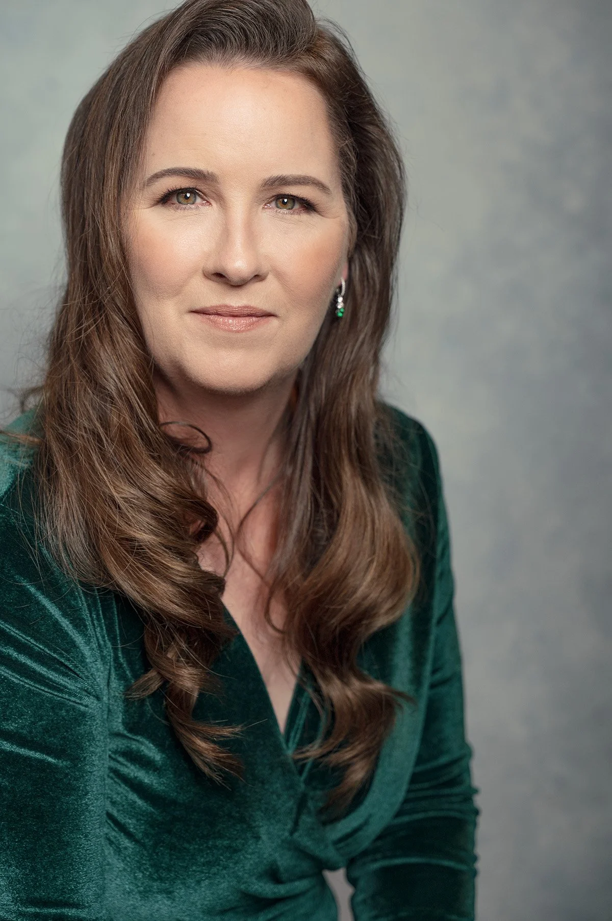 A woman with long brown hair, wearing a green velvet blazer, looking at the camera with a neutral expression, against a plain gray background in this glamorous headshot taken by Red Deer photographer.