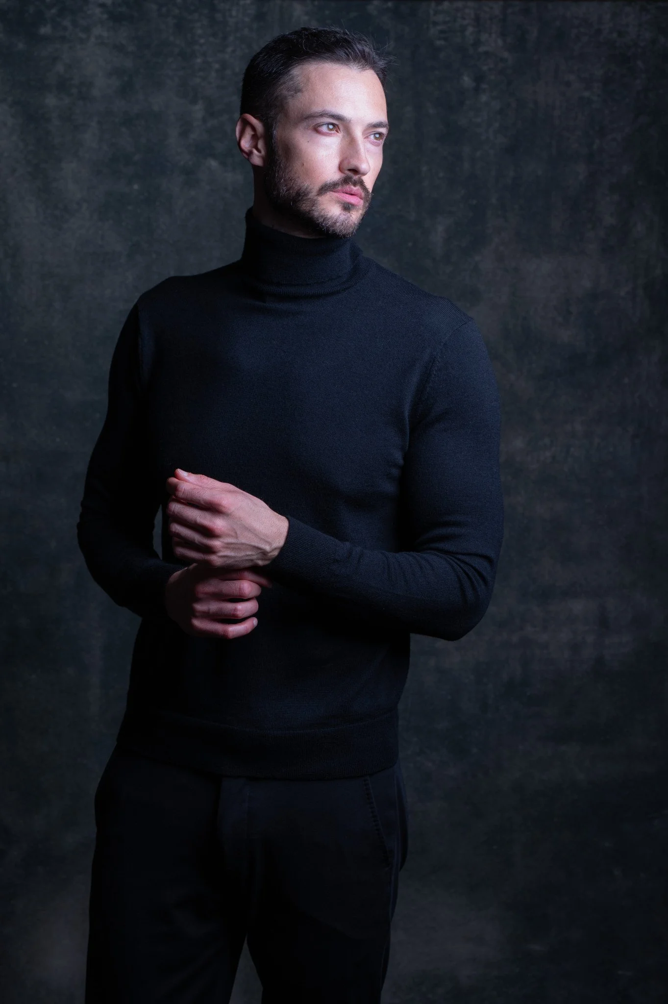 A man with dark hair, a beard, and light skin wearing a black turtleneck sweater against a dark textured background.