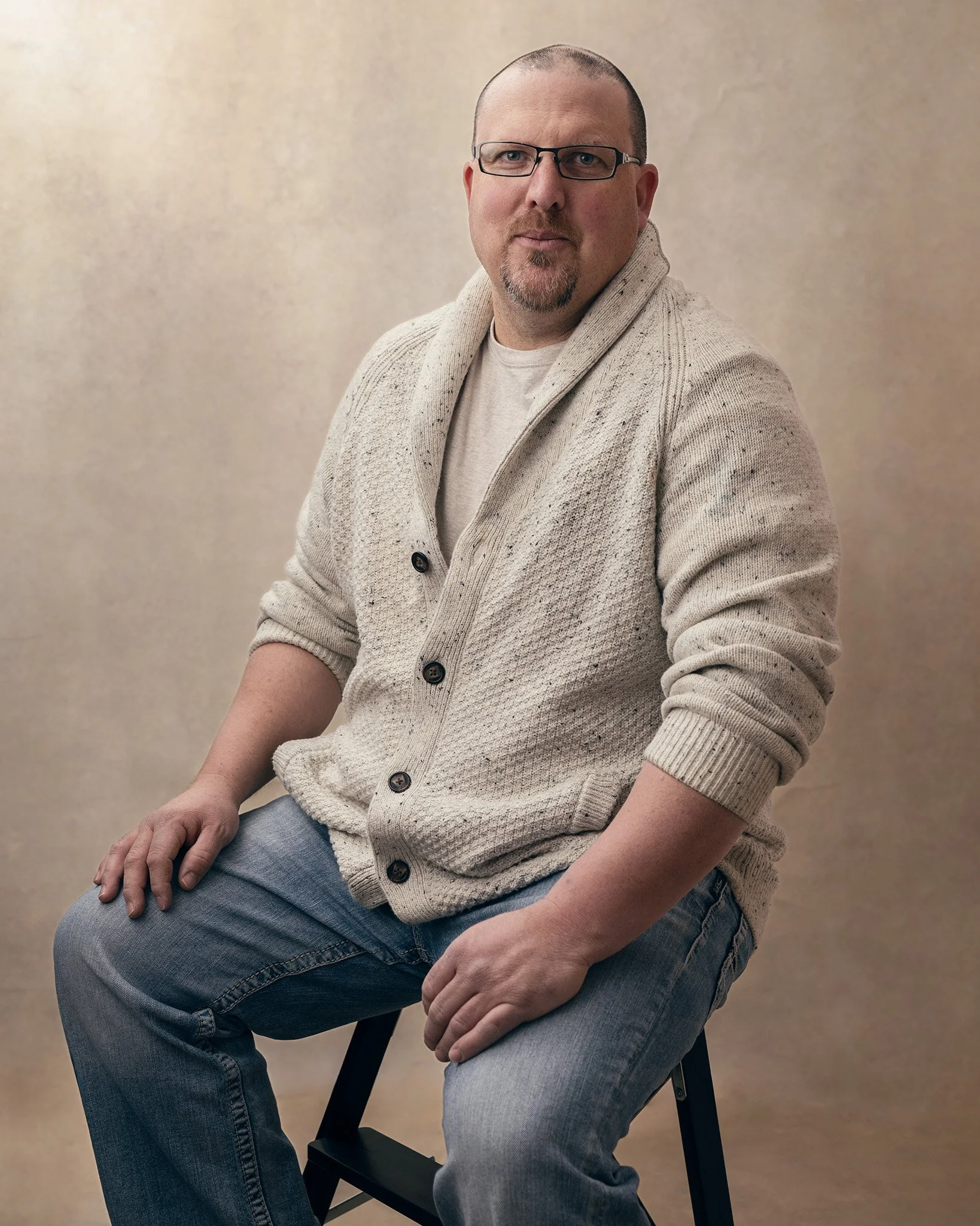 A man with glasses and a goatee wearing a beige cardigan and gray jeans, seated on a black chair against a beige background.