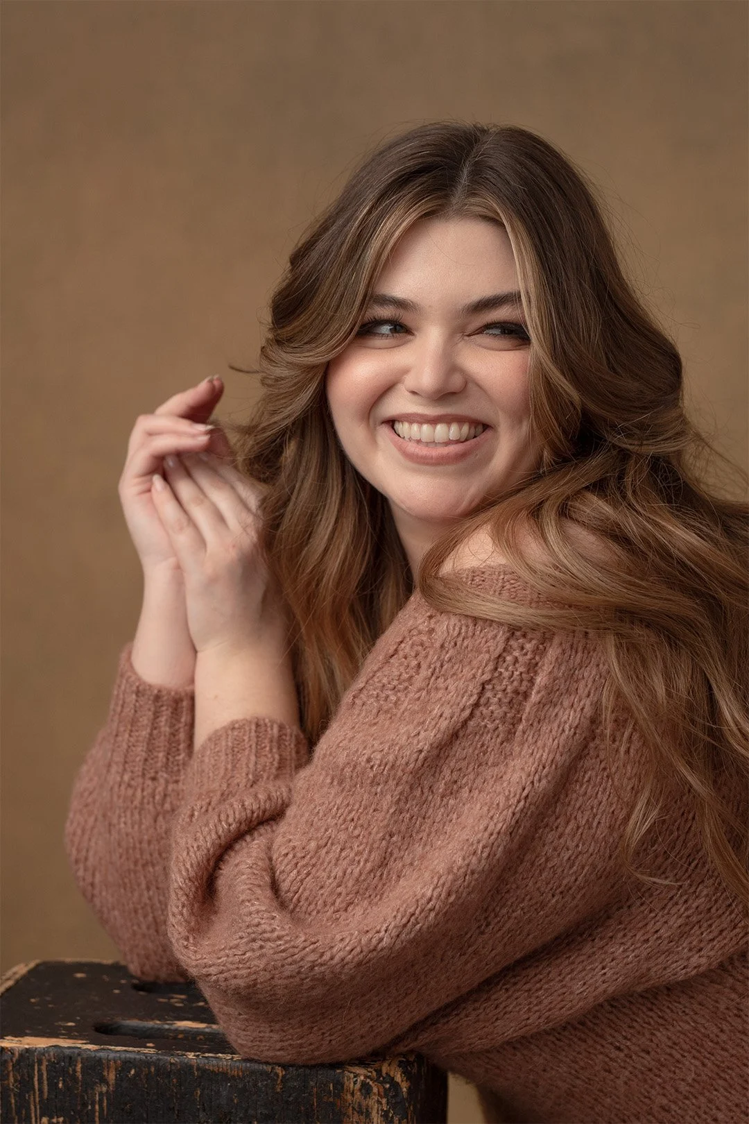 A young woman with wavy brown hair, smiling, wearing a pink knitted sweater, sitting with her elbows on a wooden surface against a brown background.