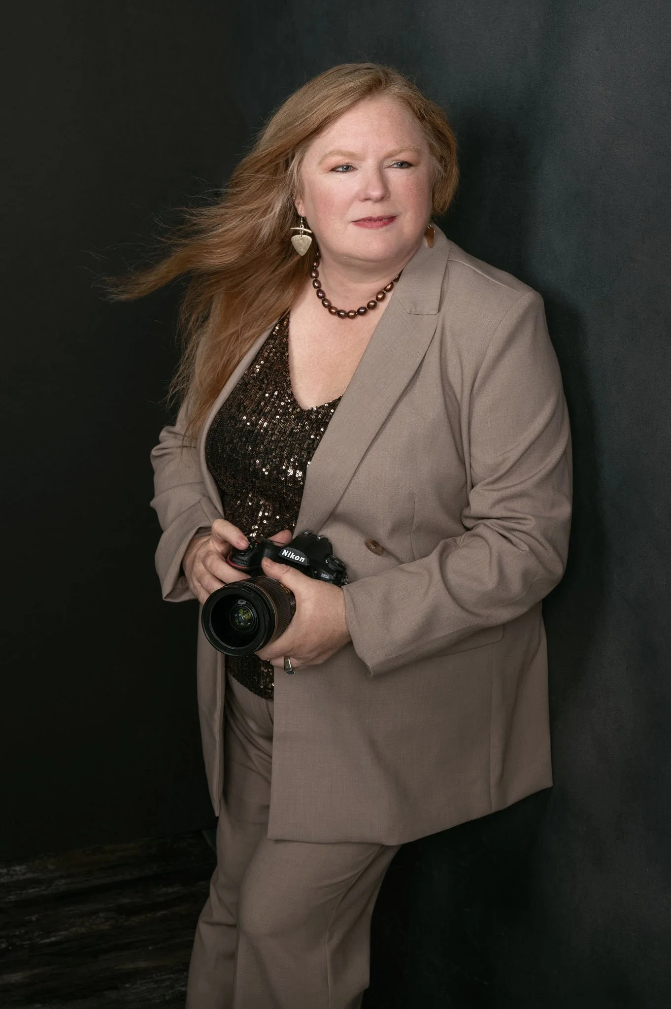 A woman with long red hair, wearing a beige suit and a black sequined top, holding a Nikon camera, standing against a dark background.