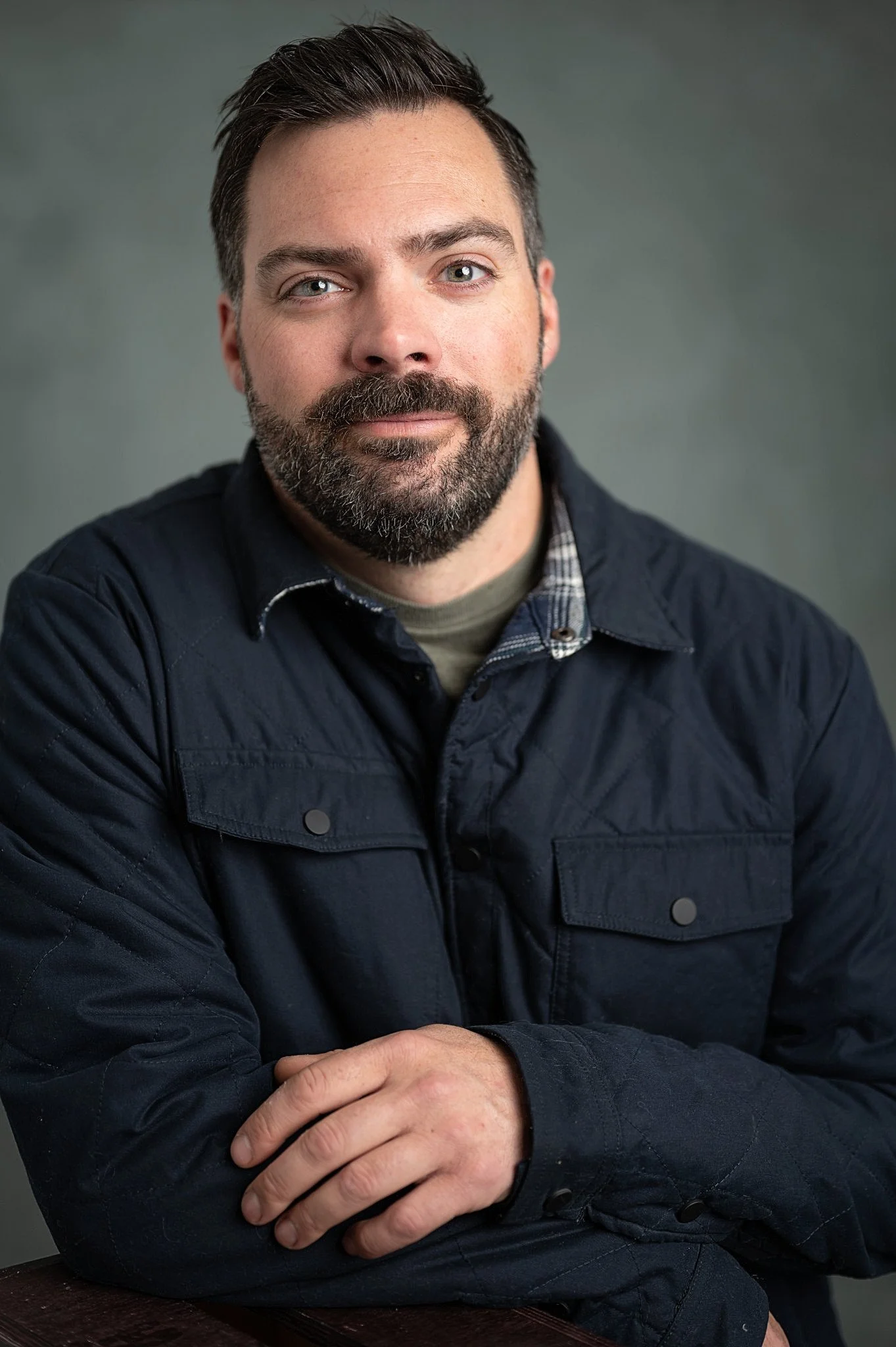 Headshot of a man with dark hair and a beard, wearing a dark jacket and a plaid shirt, against a plain background.