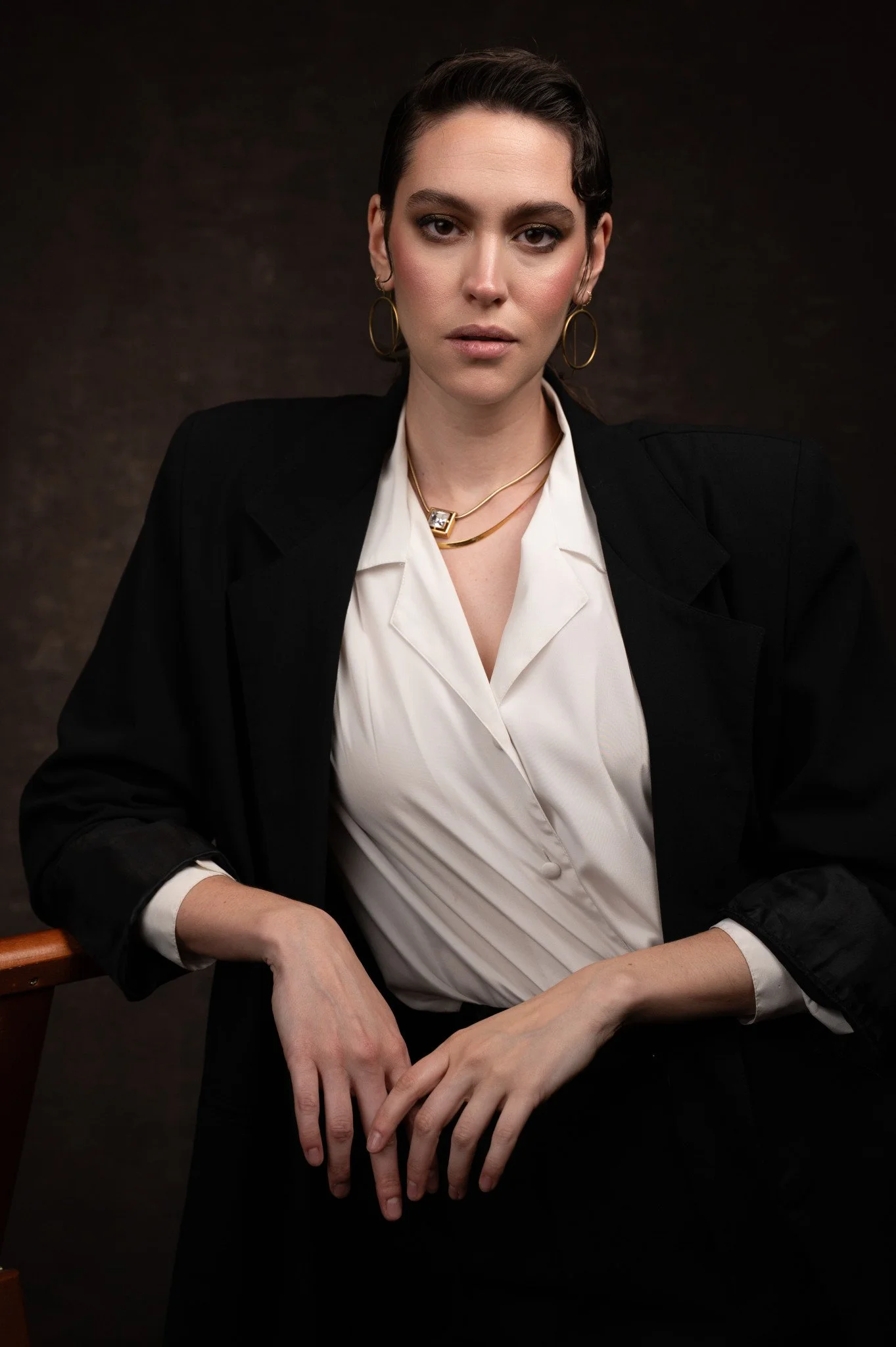A woman with short dark hair and a serious expression, wearing a black blazer over a white blouse, accessorized with gold jewelry, seated against a dark background.