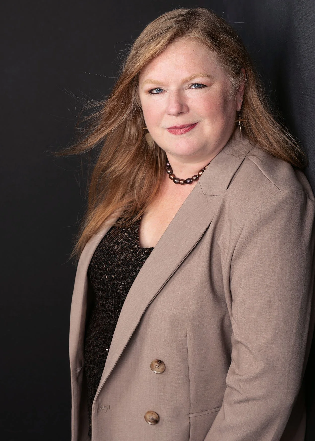 A woman with long reddish-blonde hair, wearing a beige blazer over a black top with sequins, accessorized with a pearl necklace and earrings, poses against a dark background in this professional branding image.