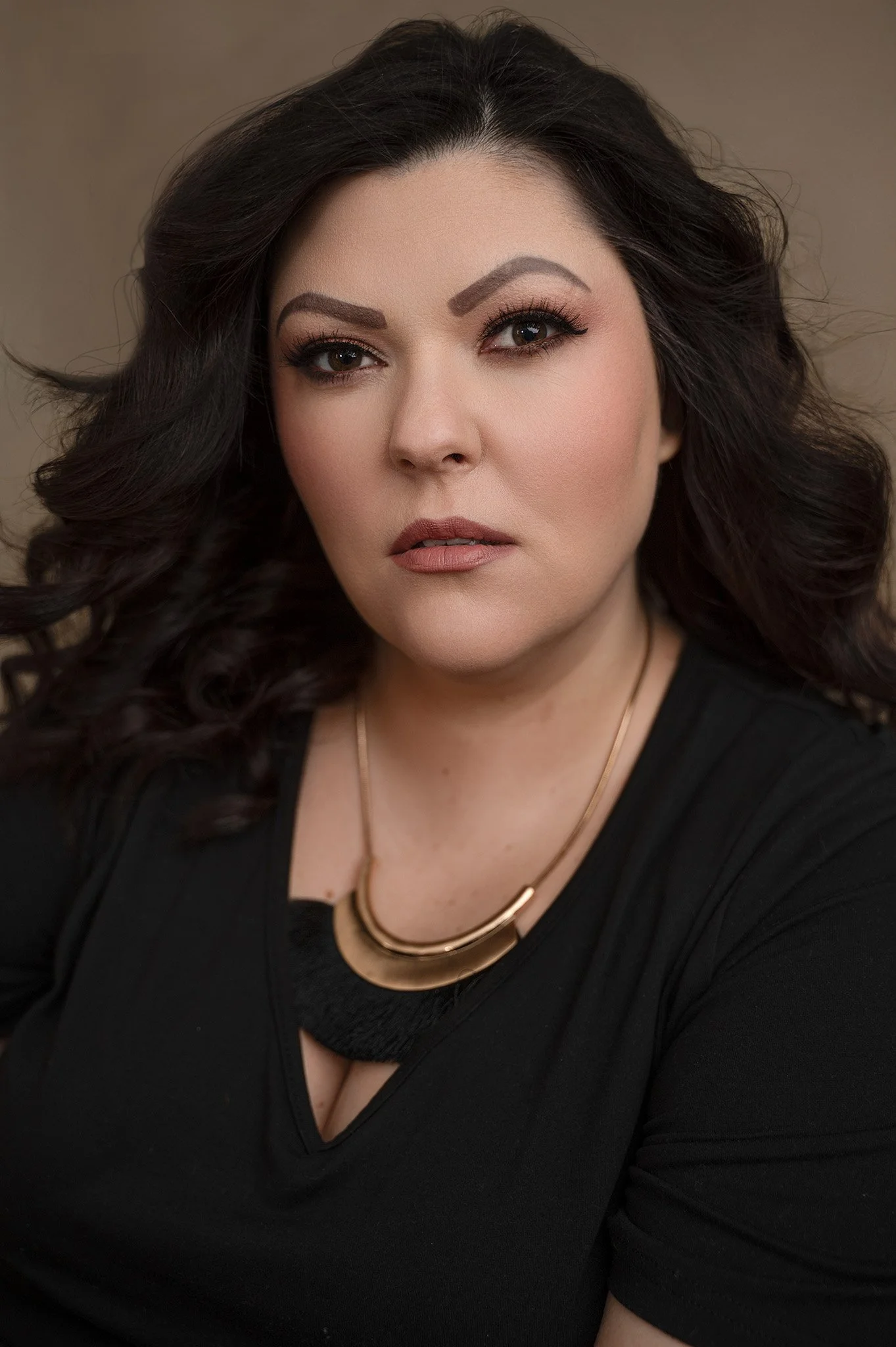 Headshot of a woman with dark wavy hair, wearing makeup, a black top, and gold jewelry, looking directly at the camera.