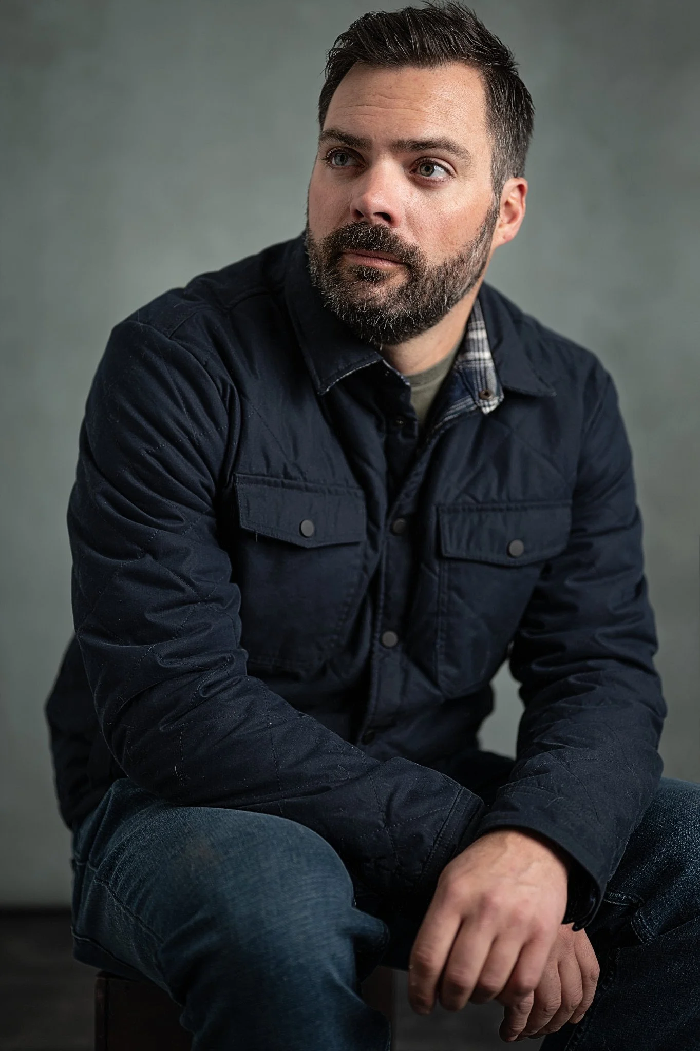 A man with a beard and short dark hair sitting against a plain gray background, wearing a dark navy jacket and jeans, looking to the side.