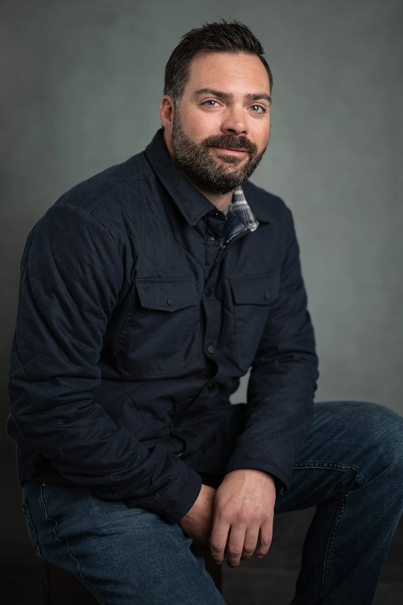 A man with dark hair and a beard, wearing a dark navy jacket and jeans, posing seated against a plain gray background.